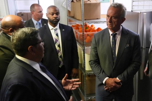 A photo of Robert F. Kennedy Jr. receiving a tour of a food distribution center. Shelves behind him show crates filled with orange bell peppers.