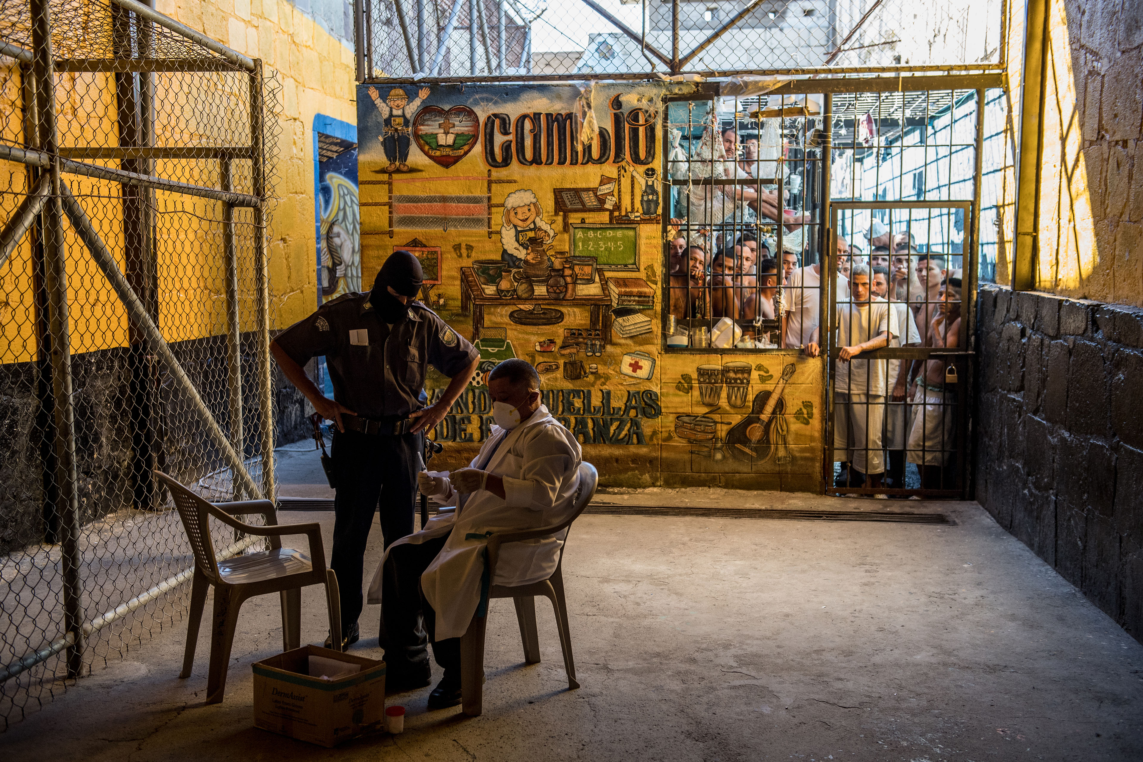 At a prison in El Salvador, men are contained behind a brightly painted wall and bar doors. In the foreground, an officer is standing with his hands on his hips as he looks over a medic who is sitting in a chair, looking at a test for tuberculosis.