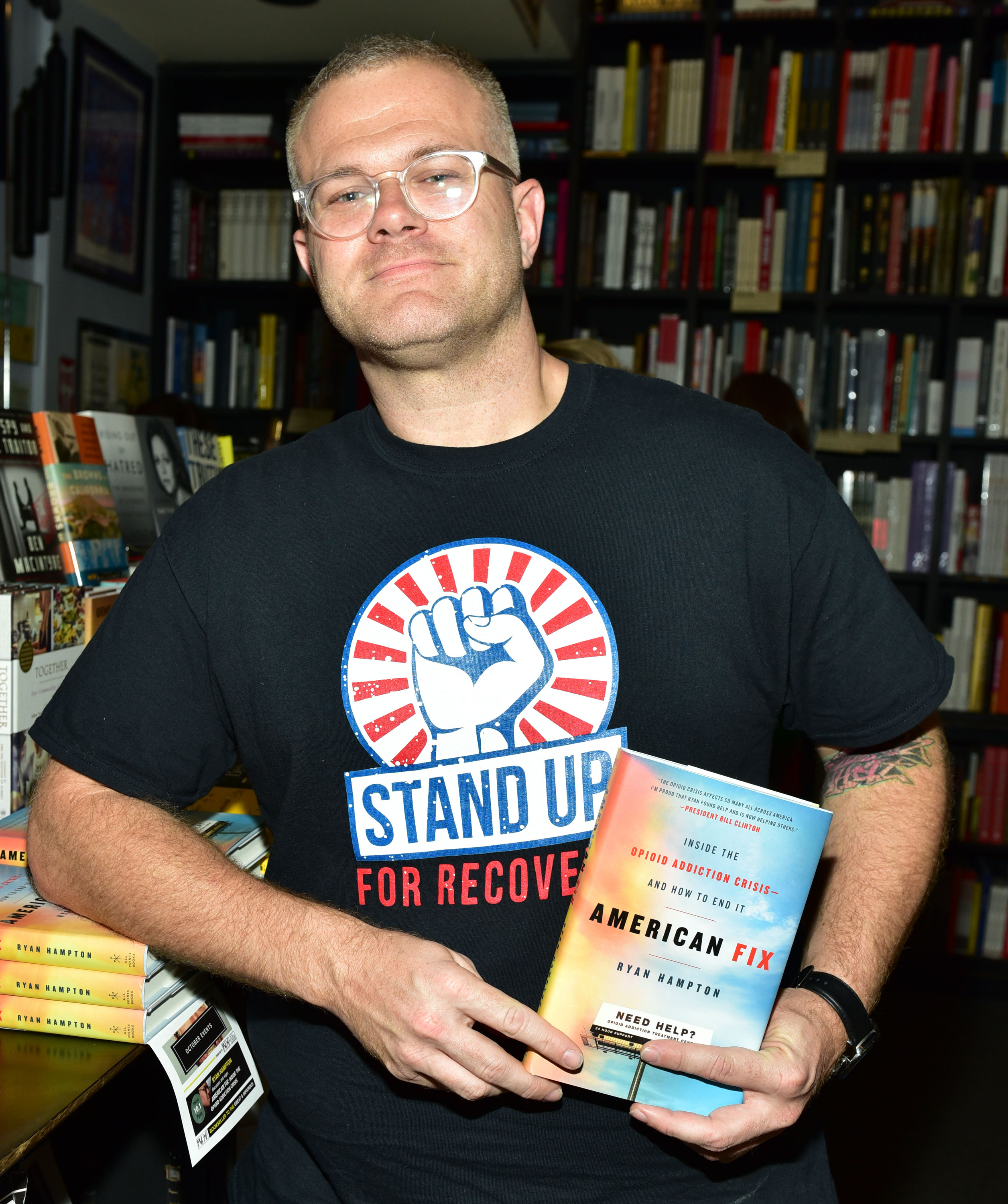 A man wearing glasses and black shirt with a logo and the words "Stand up for recovery", leans against a table display of books and holds a book titled "American Fix: Inside the Opioid Addiction Crisis and How To End It".