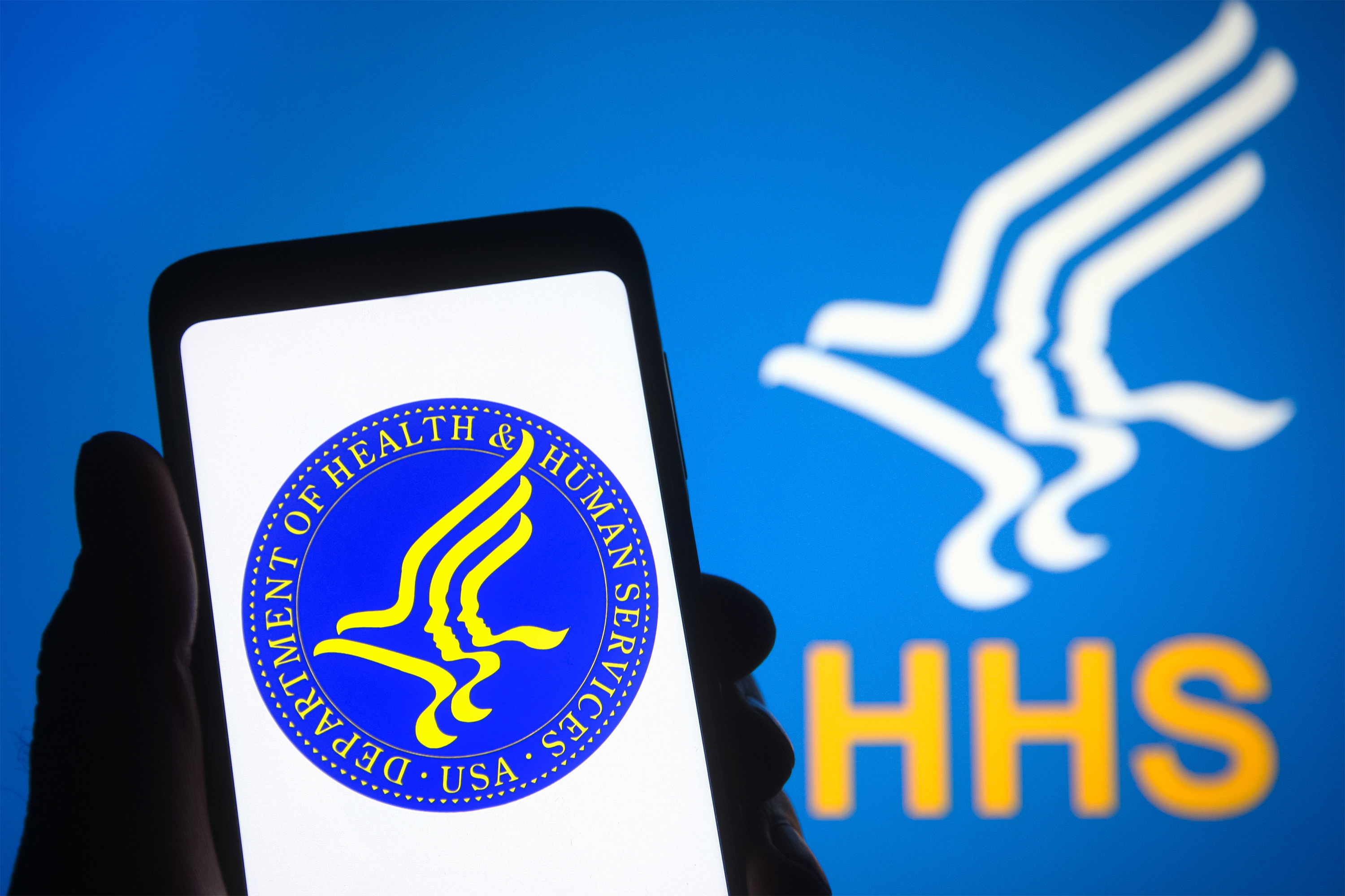 A photo of someone holding up an image on their phone of the Department of Health and Human Services logo. Behind them is a backdrop with the same logo.