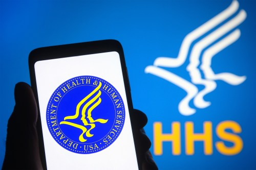 A photo of someone holding up an image on their phone of the Department of Health and Human Services logo. Behind them is a backdrop with the same logo.