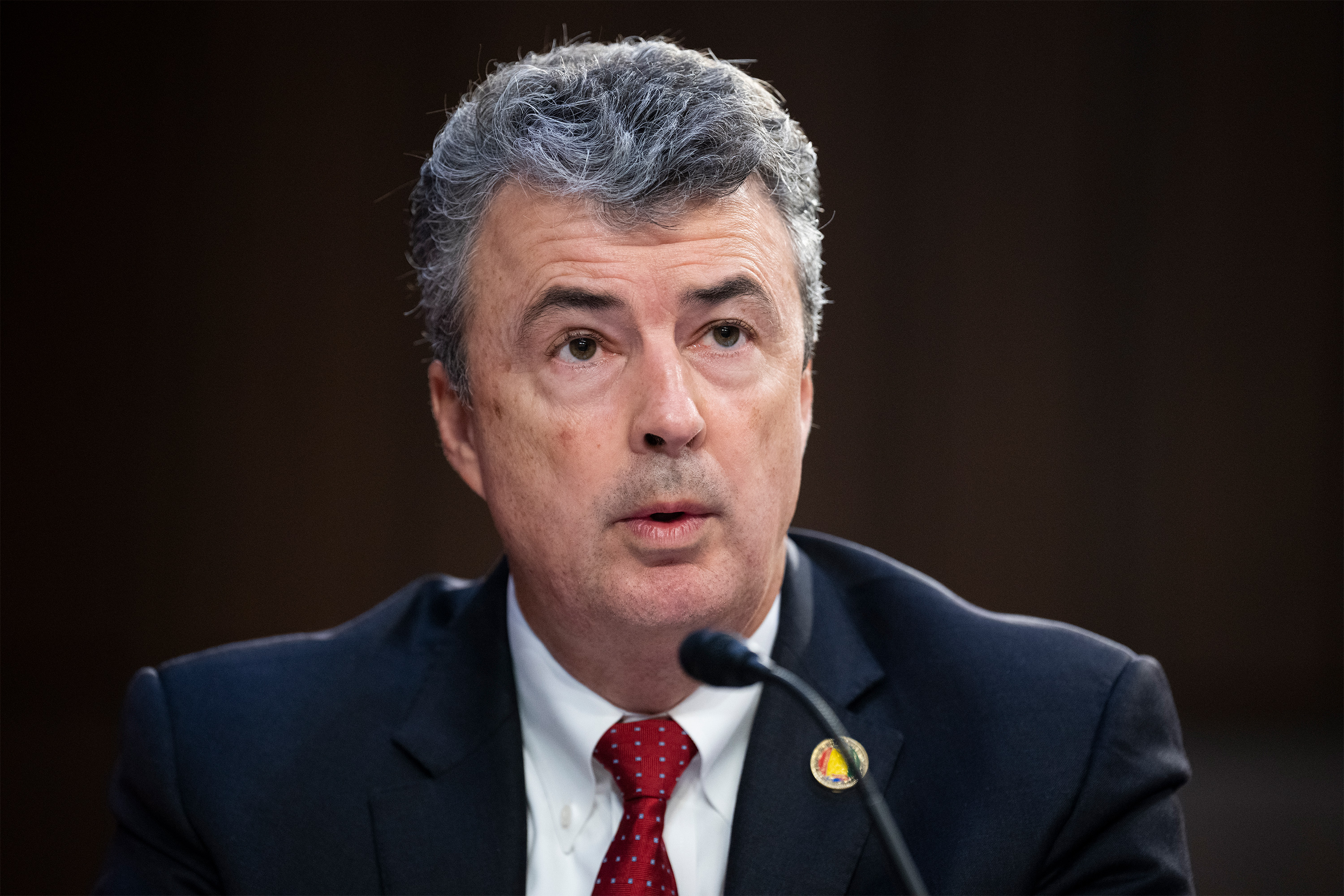 A photo of a man sitting in a Senate hearing room, speaking into a microphone at a desk.