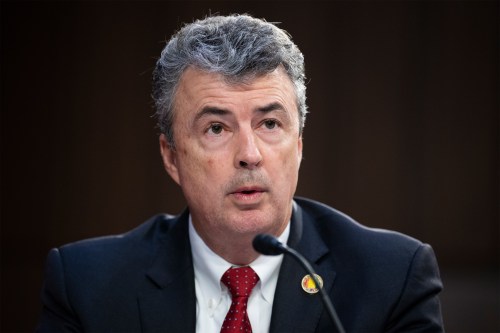 A photo of a man sitting in a Senate hearing room, speaking into a microphone at a desk.