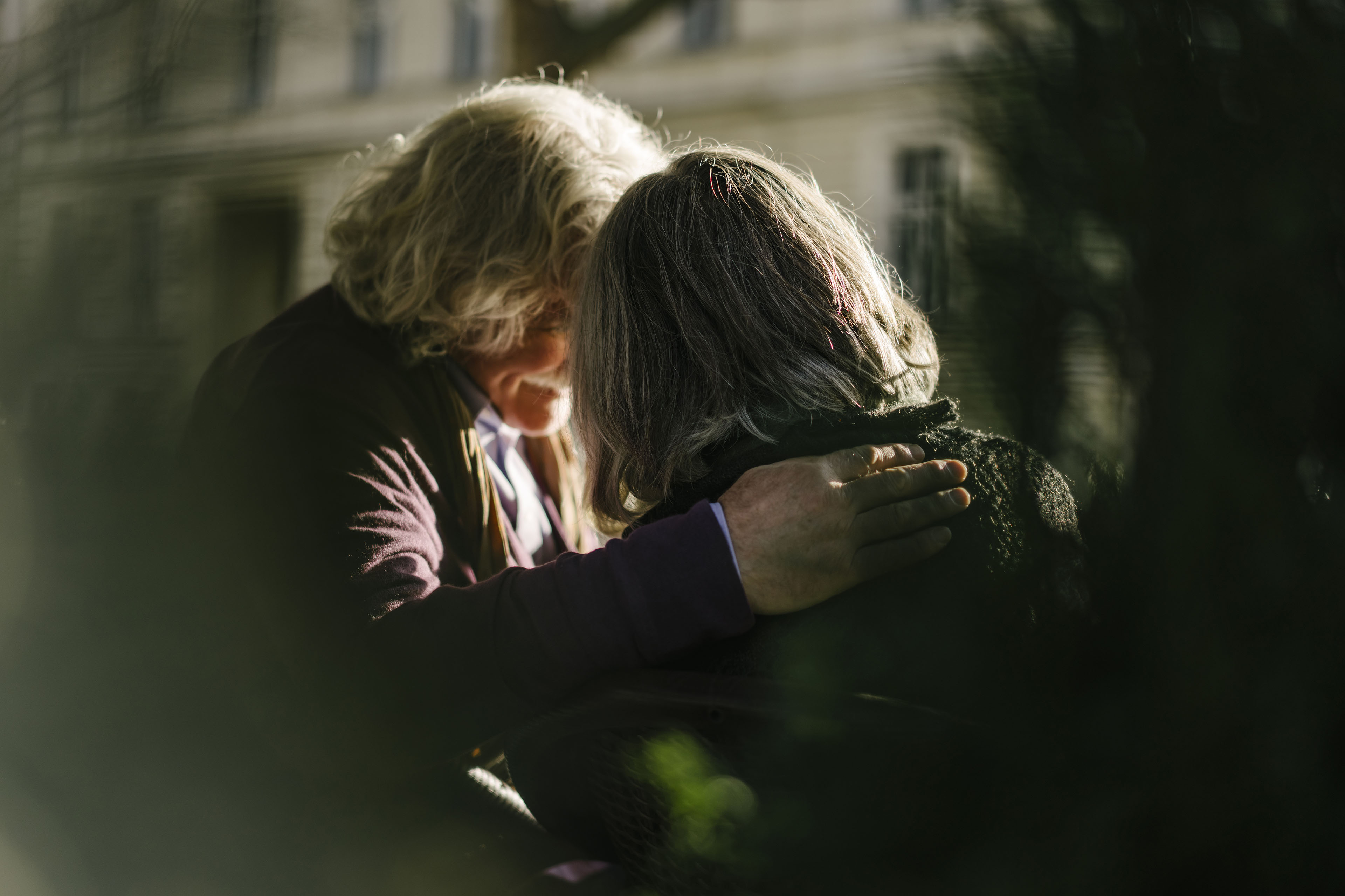 An unidentifiable grey haired senior couple are photographed from behind, their faces obscured from view. One has their hand on the other's shoulder.