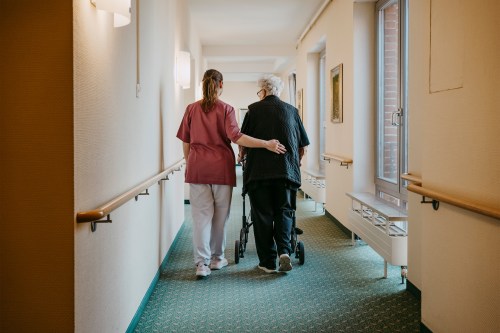 A photo of female caregiver escorting an elderly woman patient down a hallway in a nursing home.