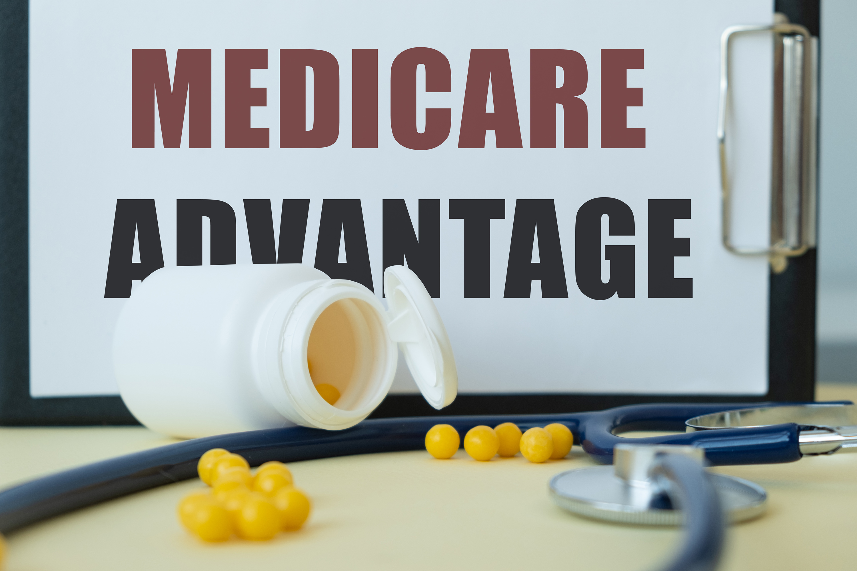 A photo of a clipboard resting horizontally on a table with a piece of paper that reads, "Medicare Advantage." In front of it are an open pill bottle spilled yellow capsules, a stethoscope, and a pen.