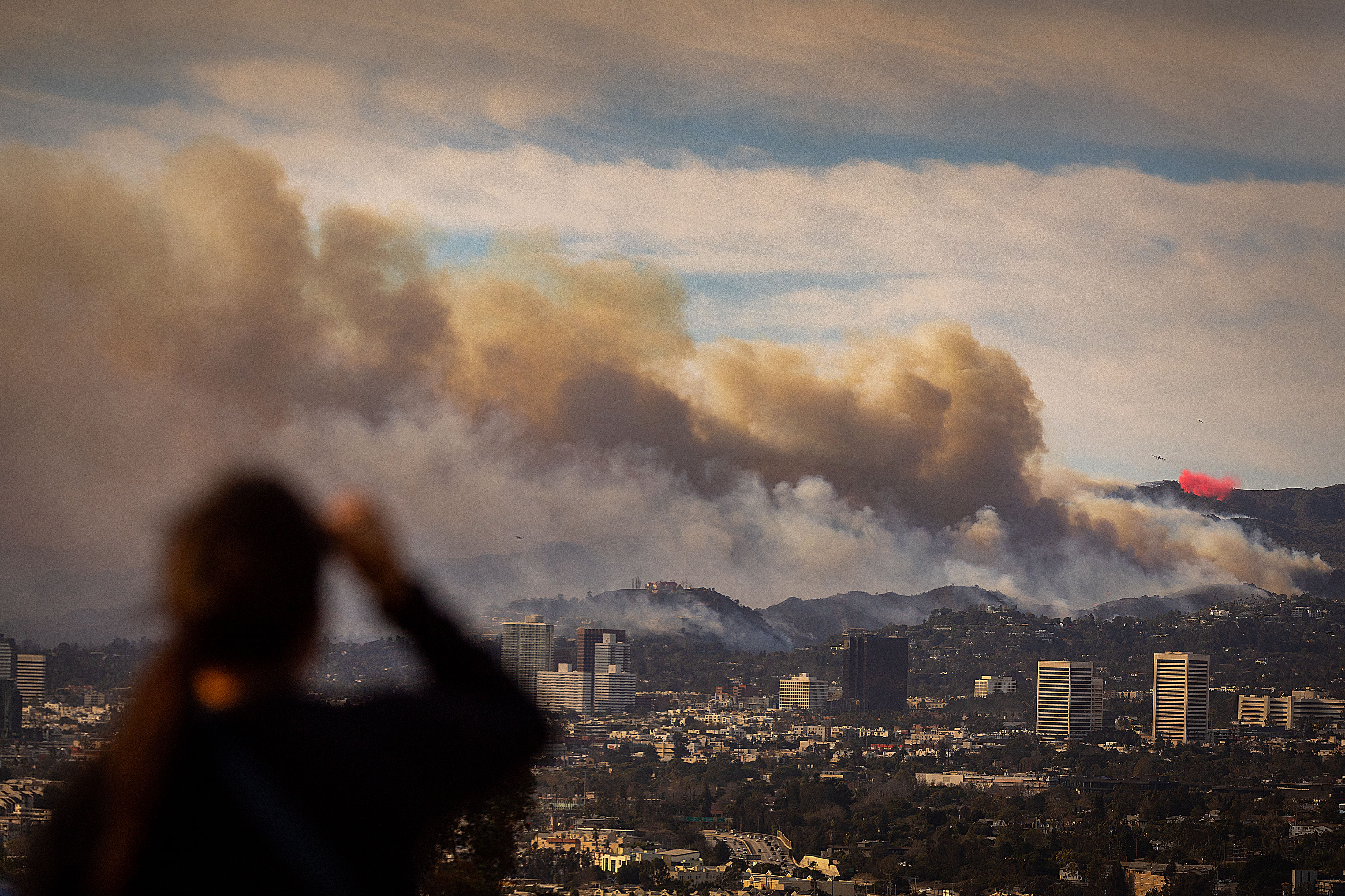 A person is out of focus on a hill in the foreground looking out at Los Angeles and billowing smoke from wildfires obscuring the view.
