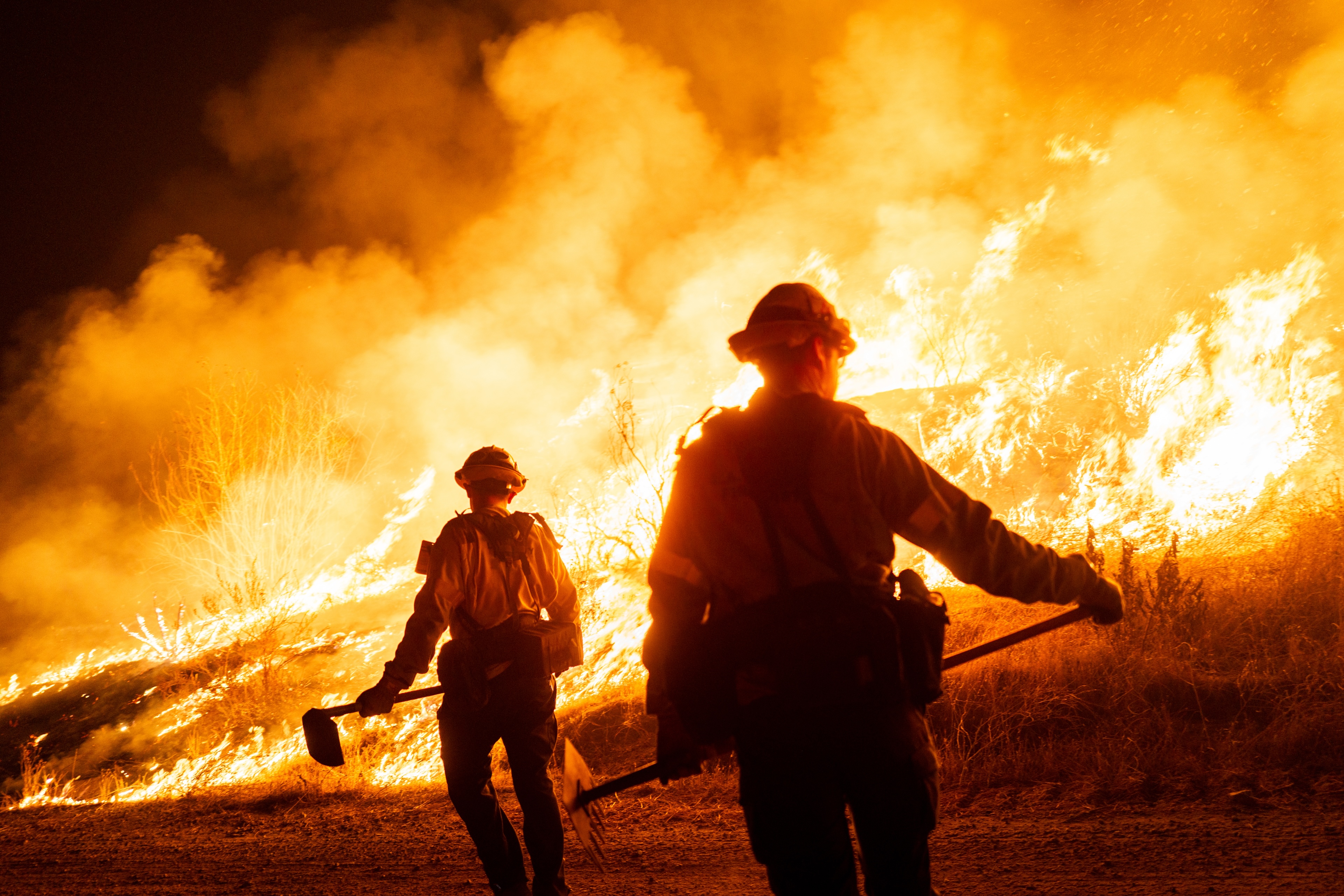 firefighters with shovels stand before a large orange blaze and smoke plumes