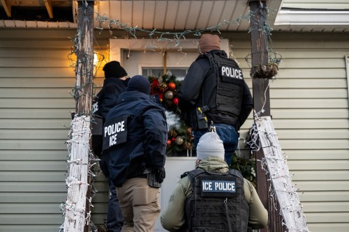US Immigration and Customs Enforcement (ICE) agents knock on the door of a residence. Three men are seen standing on the stairs of a small wooden porch, their backs to the camera.