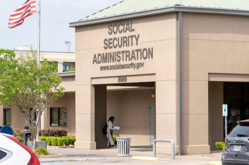 A photo of a concrete office building with the words "Social Security Administration" and "www.socialsecurity.gov" on a sign on the front entrance. An American flag on a pole is posted near the entrance.