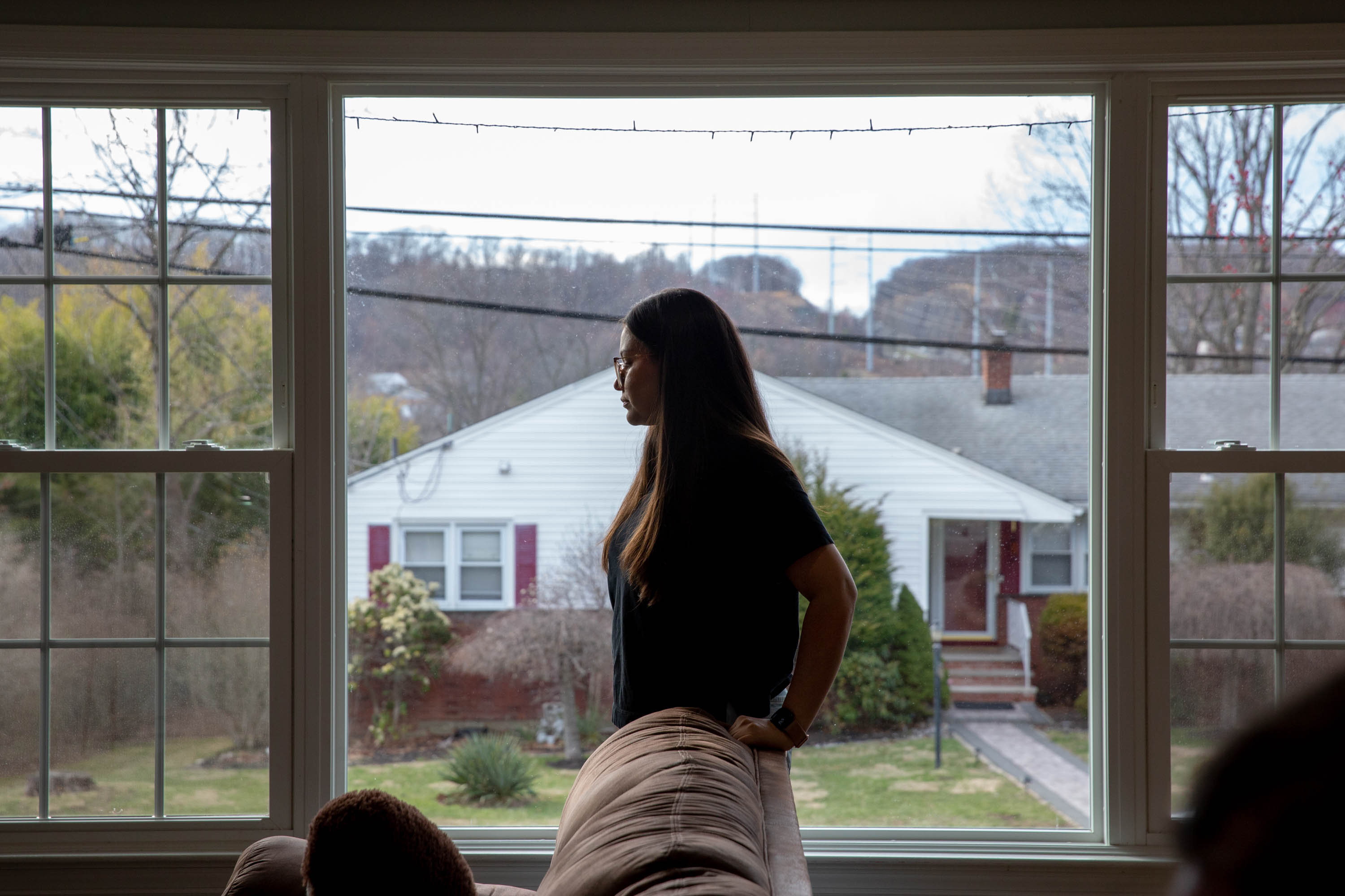 A woman with long brown hair wearing glasses stands in front of a window in a home on a suburban street