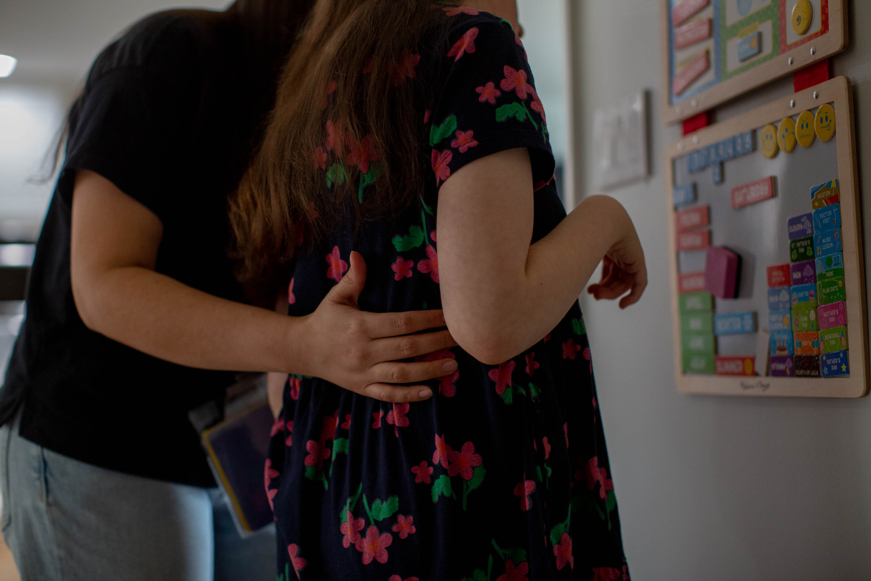 A woman in a black shirt holds her arm along the back of a girl wearing a flower dress as they look at a wall calendar together