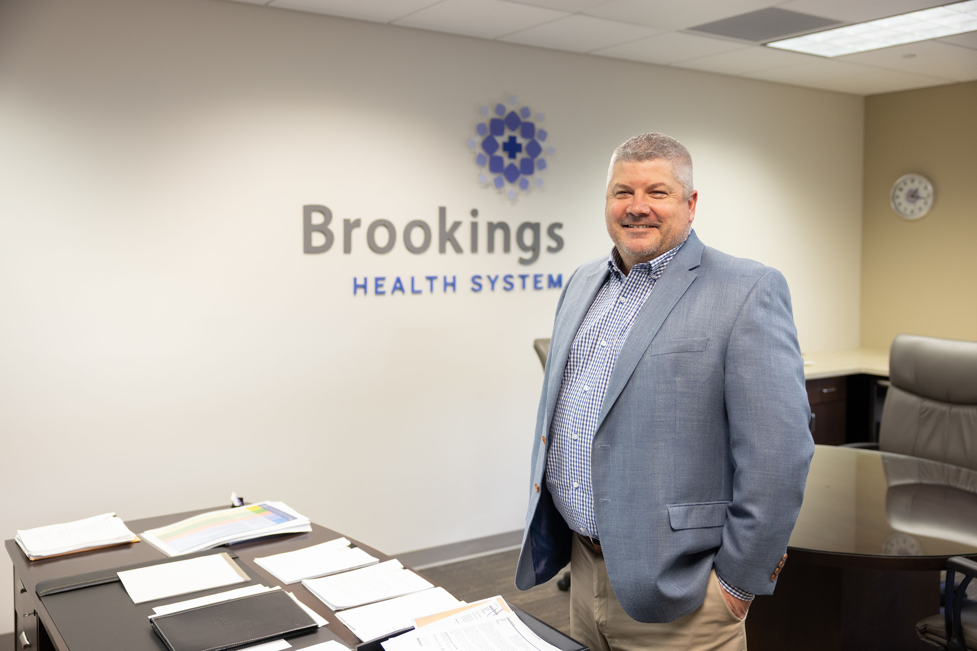 A man wearing business attire stands in an office room, smiling.