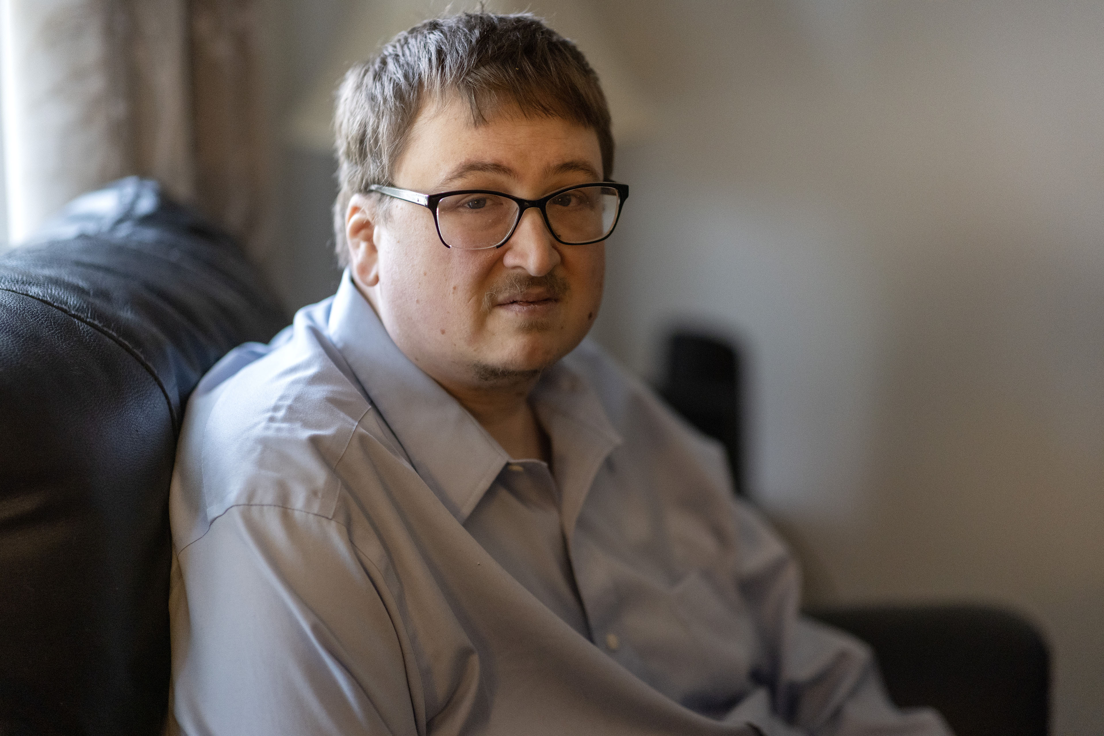 A man wearing glasses sits on a couch facing the camera.