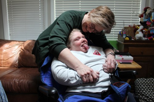 A mother leans over her child, who is in a wheelchair. They are both smiling.