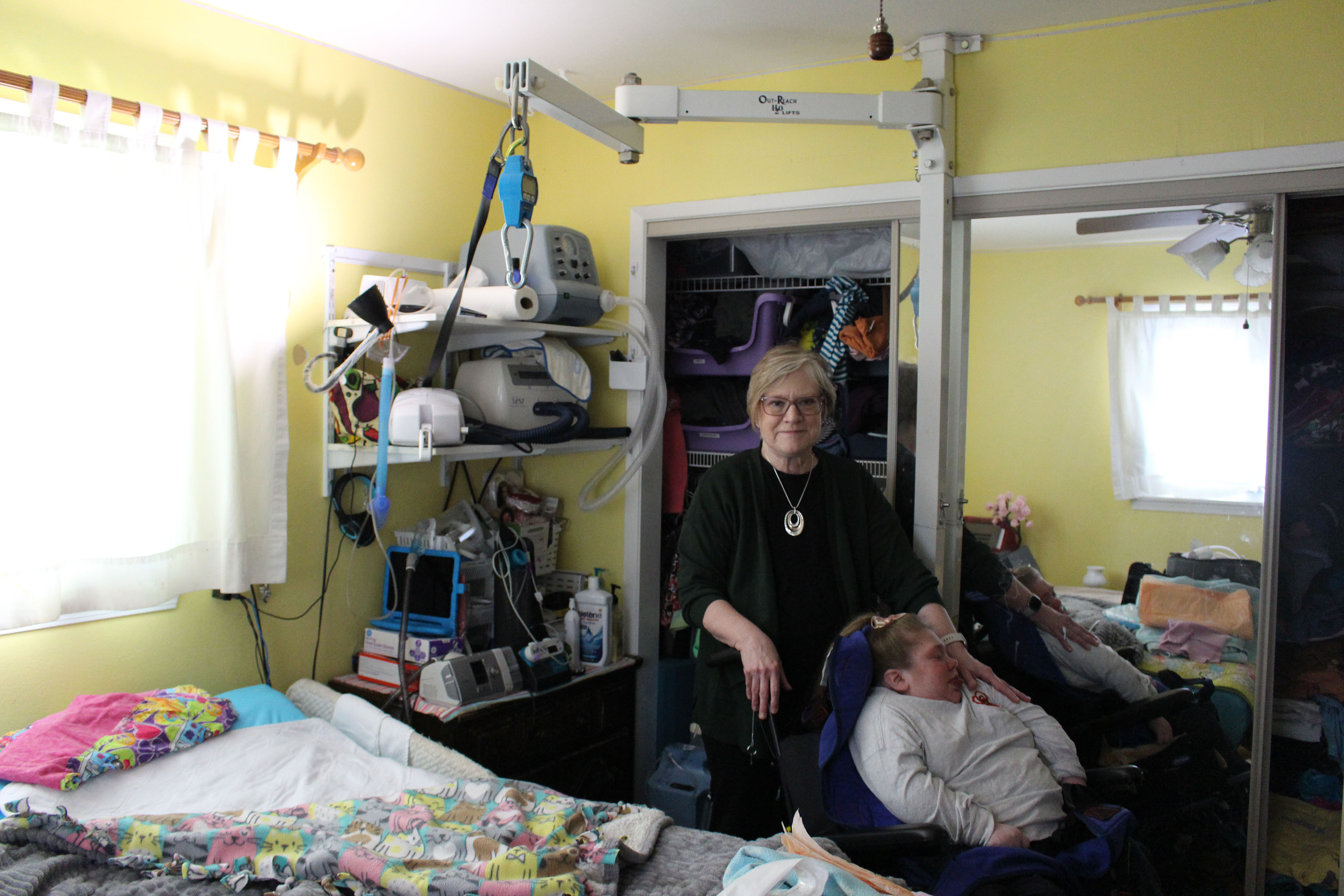 A mother stands beside her daughter, who is in a wheelchair, in her daughter's room. The room is  equipped with a device that helps lift her daughter in and out of bed, and other equipment that assists mobility and health care.
