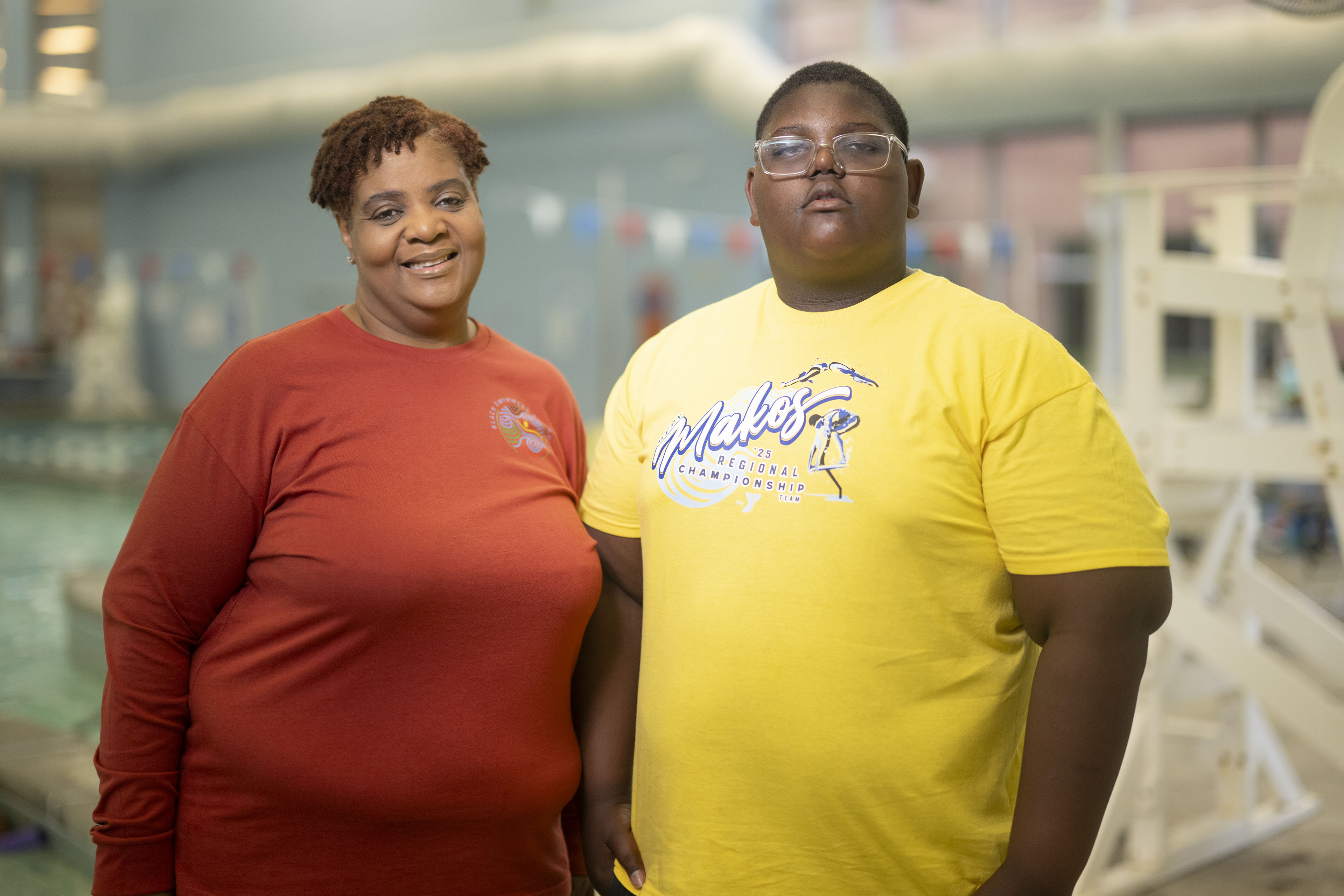 A woman wearing an orange shirt poses for a portrait in front of a pool as she stands next to a young man wearing glasses and a yellow "Makos" shirt