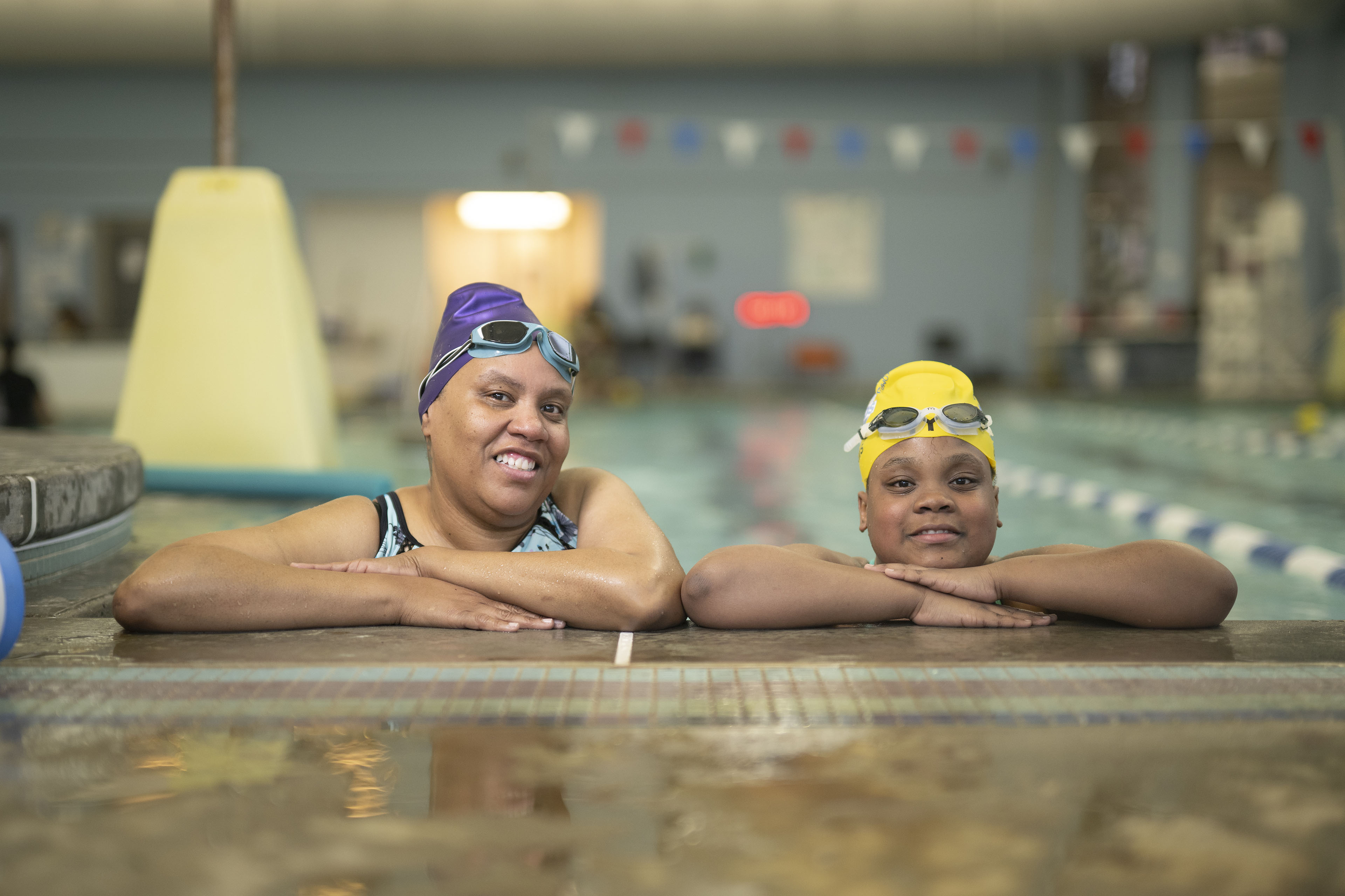 A woman wearing a purple swim cap and goggles poses at the edge of a pool with a girl wearing a yellow swim cap and goggles