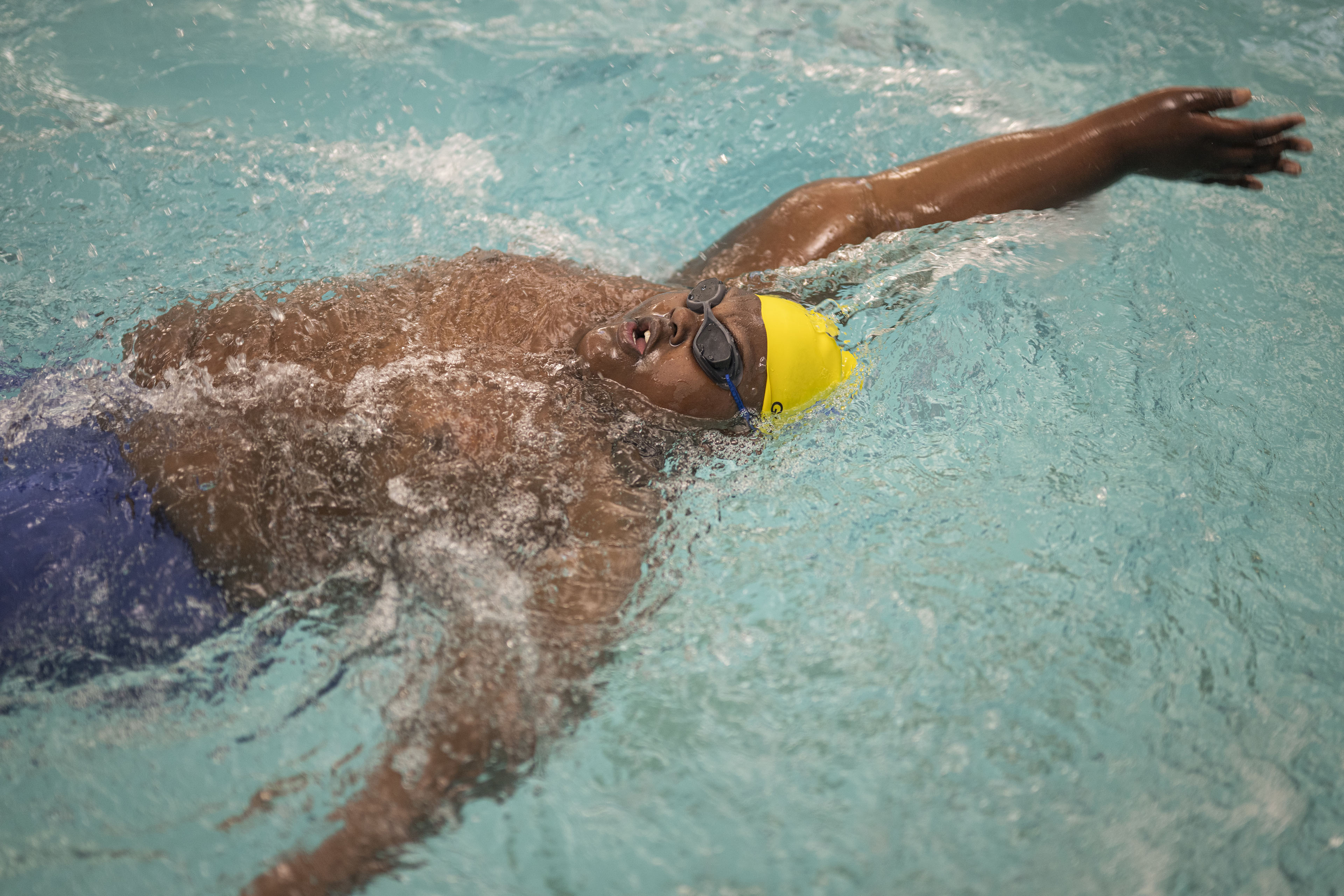 A young man wearing blue swim shorts, goggles, and a yellow swim cap does the backstroke in a pool