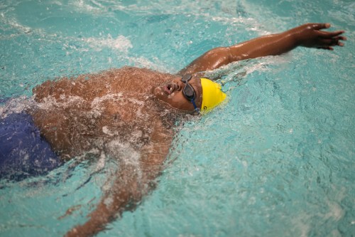 A young man wearing blue swim shorts, goggles, and a yellow swim cap does the backstroke in a pool