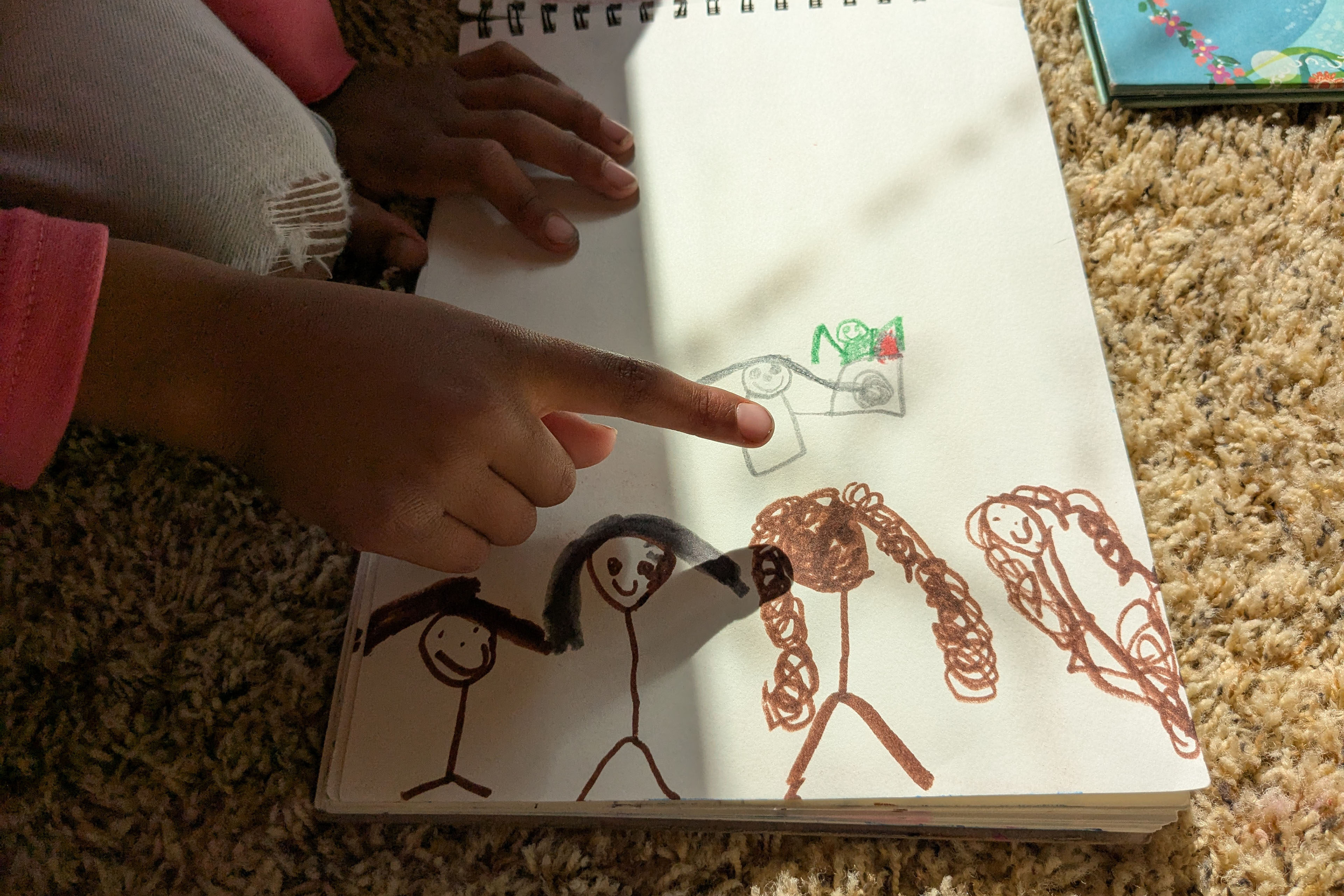 A young child points to a piece of paper. There is a drawing, made by the child, of herself and her family, as stick figures.