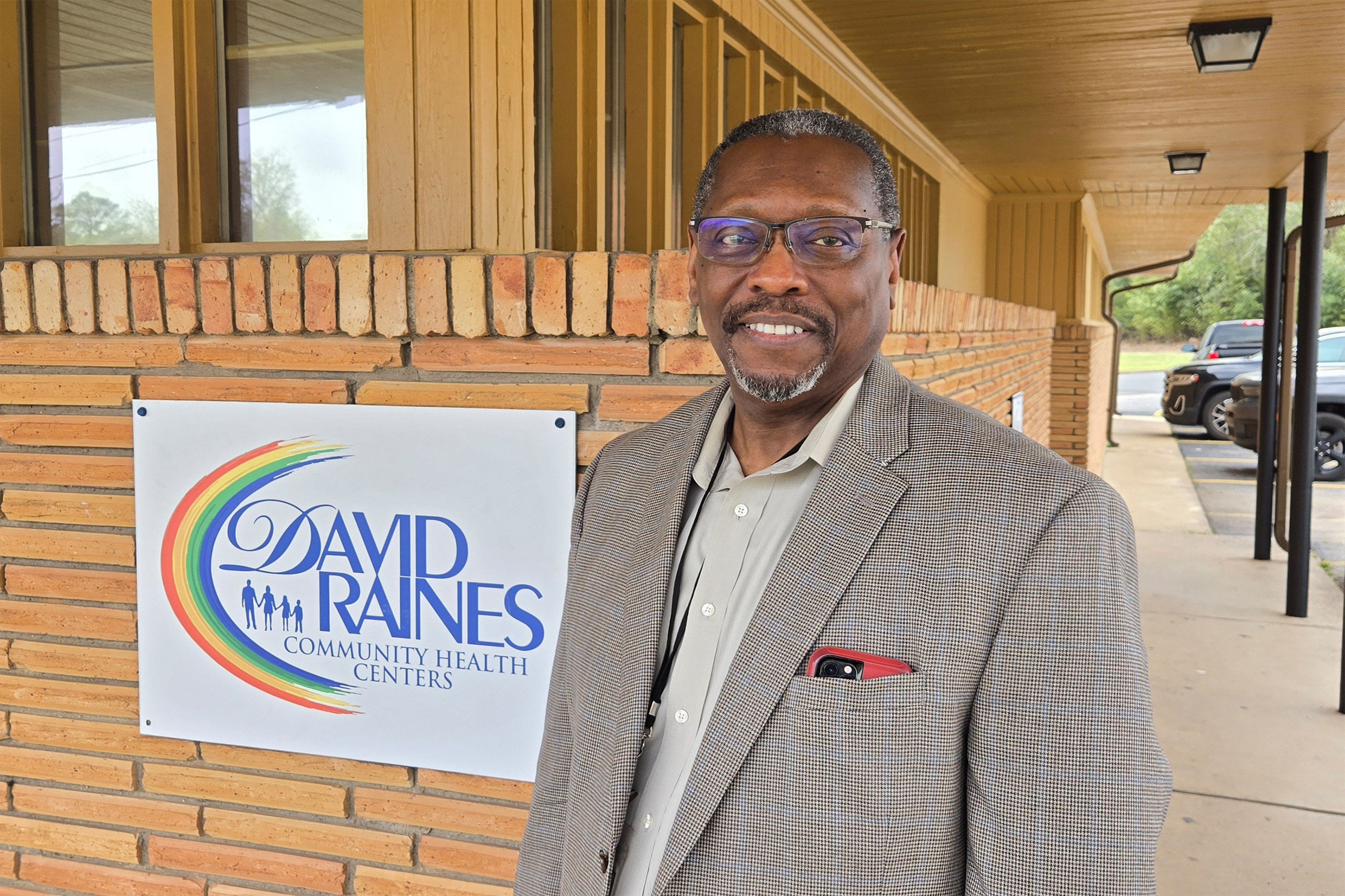 A photo of a man in a suit smiling for a photo next to a sign that reads "David Raines Community Health Centers."