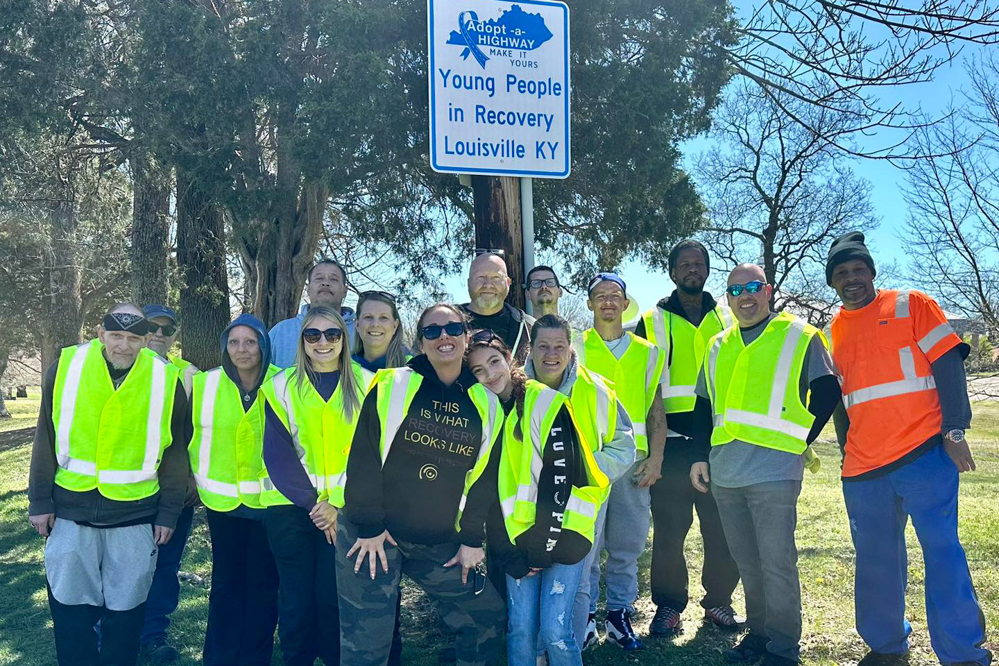 A photo of a group of people in reflective vests standing in front of a An Adopt a Highway sign that reads, "Young People in Recovery, Louisville, KY."