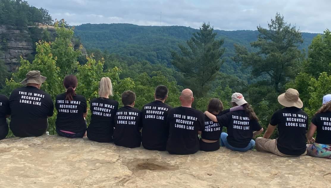 A photo of people sitting on a cliff after a hike. All of them are wearing shirts that read, "This is what recovery looks like."