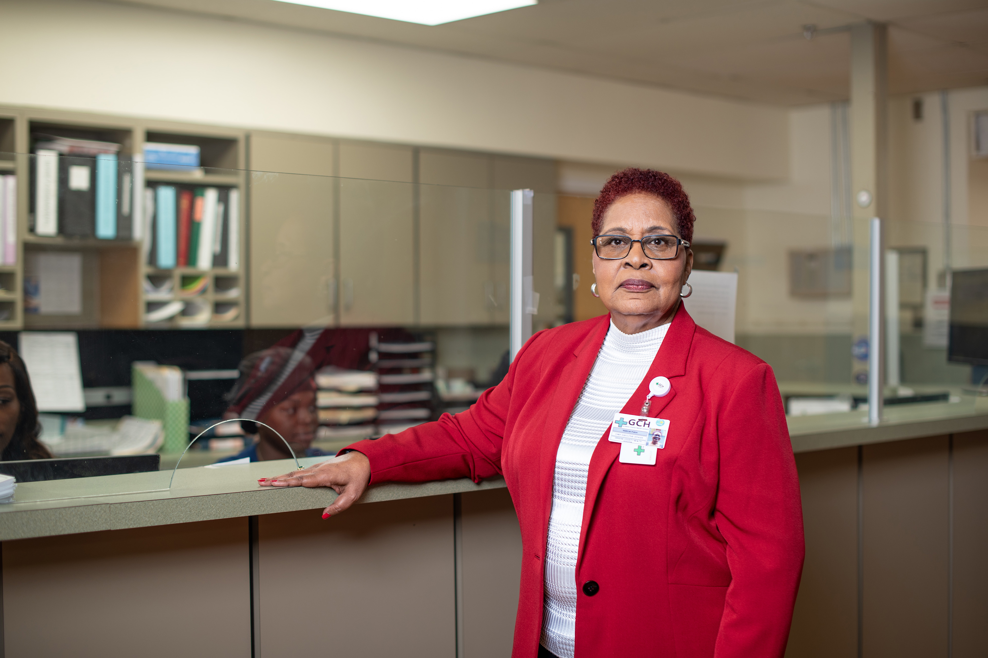 A woman wearing glasses, a white shirt, and a red blazer poses for a photo a desk at a hospital