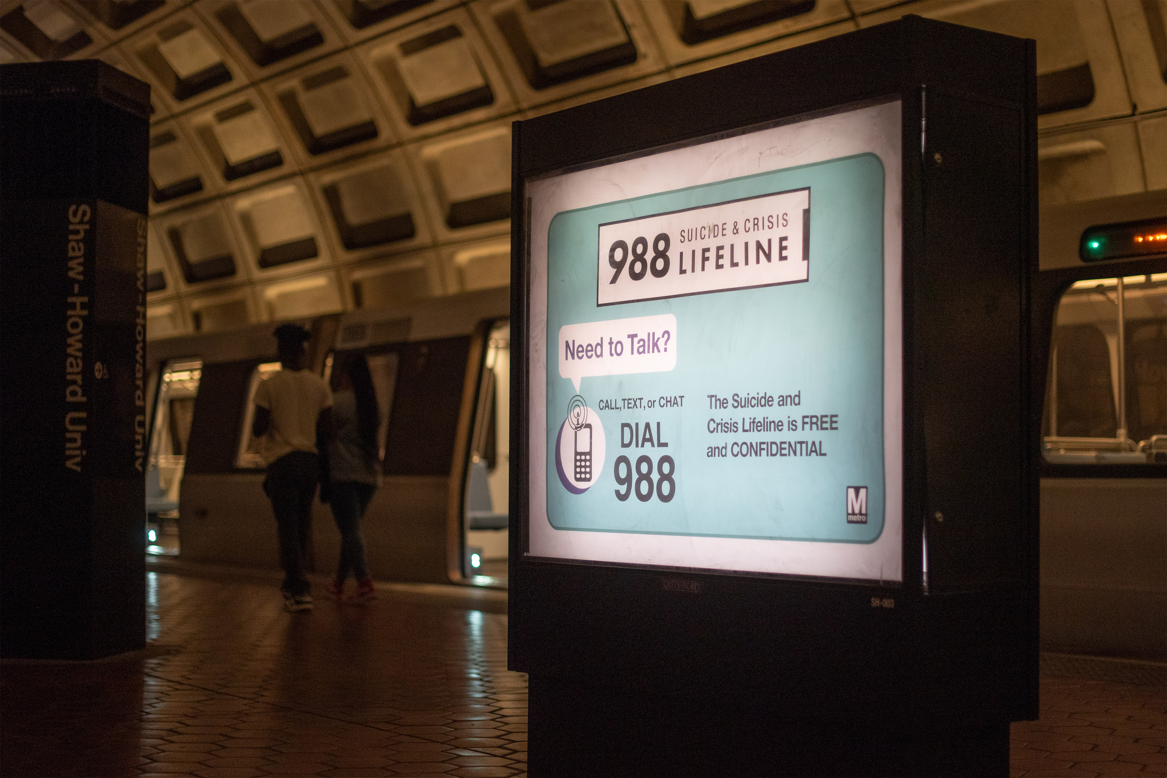 A sign on a subway platform reads, "Need to talk? Dial 988. The Suicide and Crisis Lifeline is free and confidential."