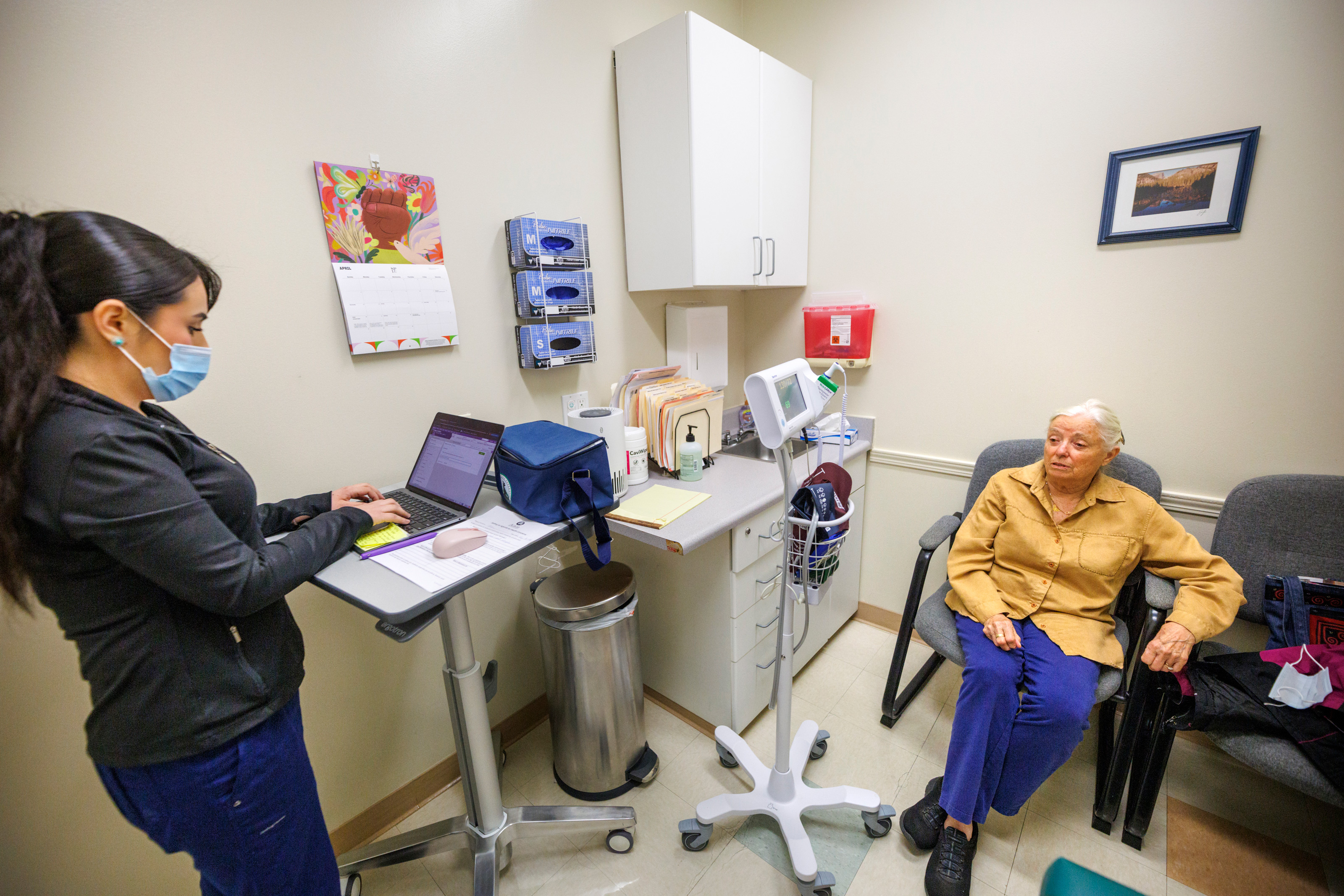 A photo of a woman speaking to a patient in an exam room while typing on a computer.