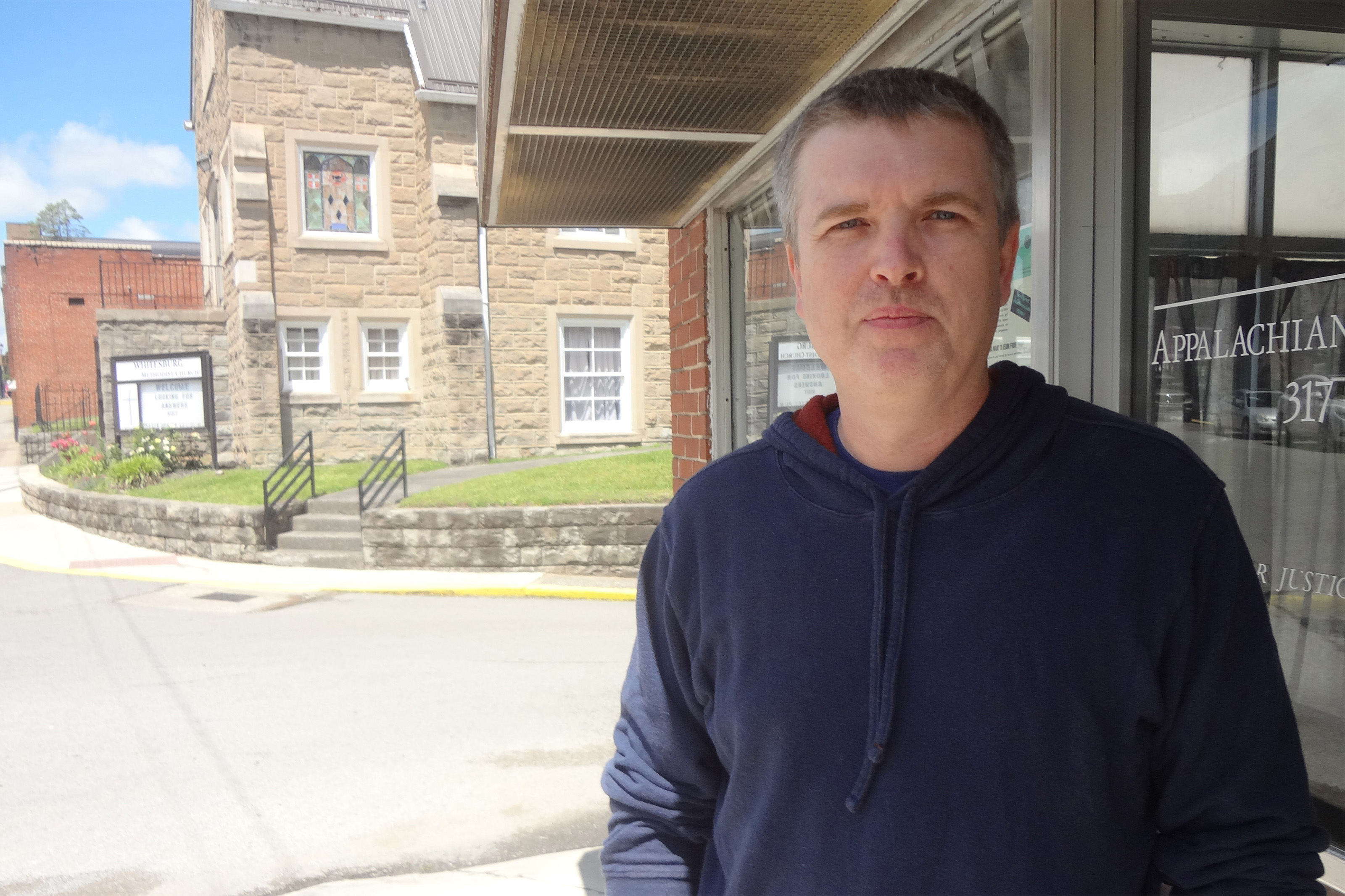 A photo of a man standing under a shaded overhang outside in front of an office building.