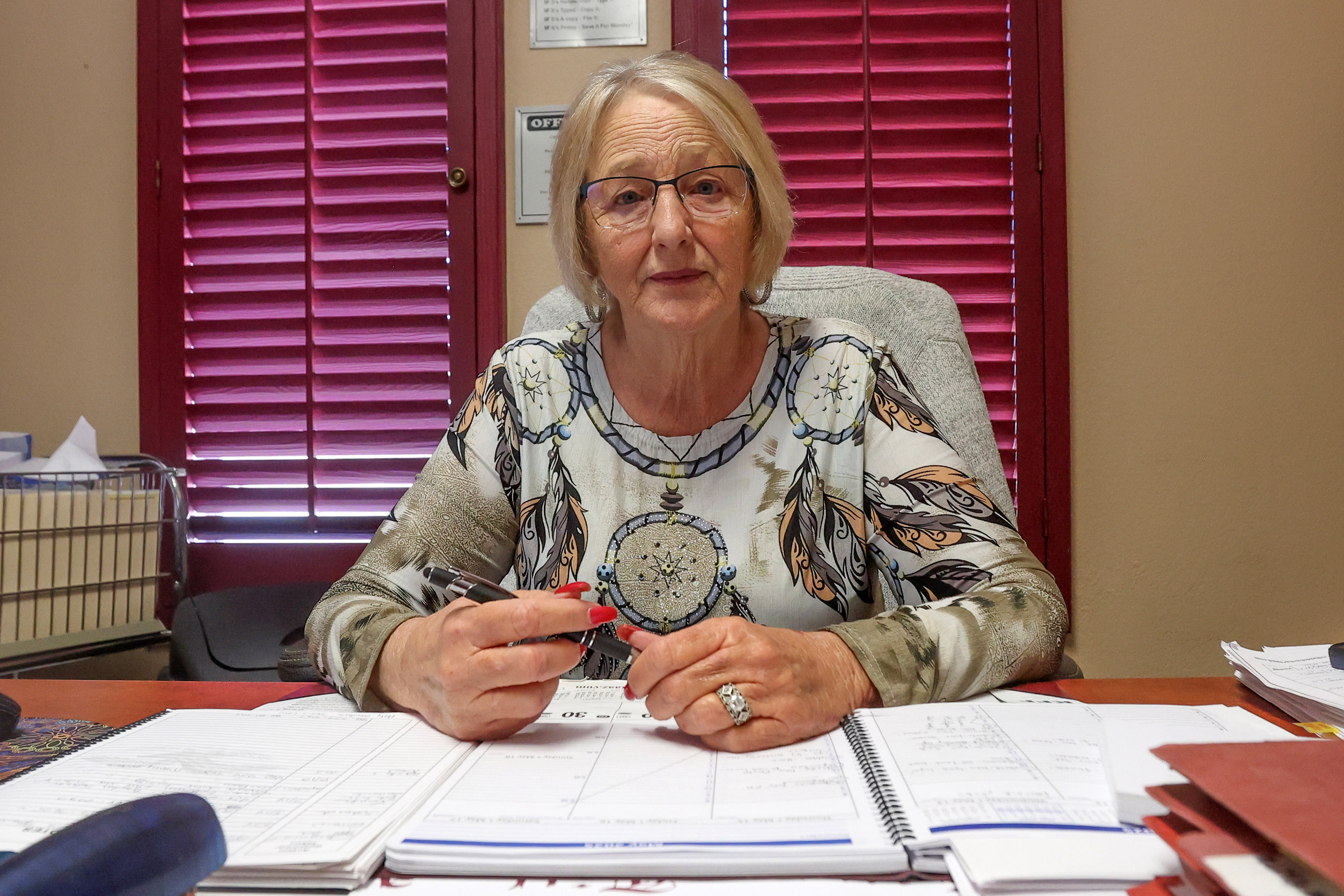A photo of a woman seated at her desk. Paperwork is in front of her and she is holding a pen in her hands.