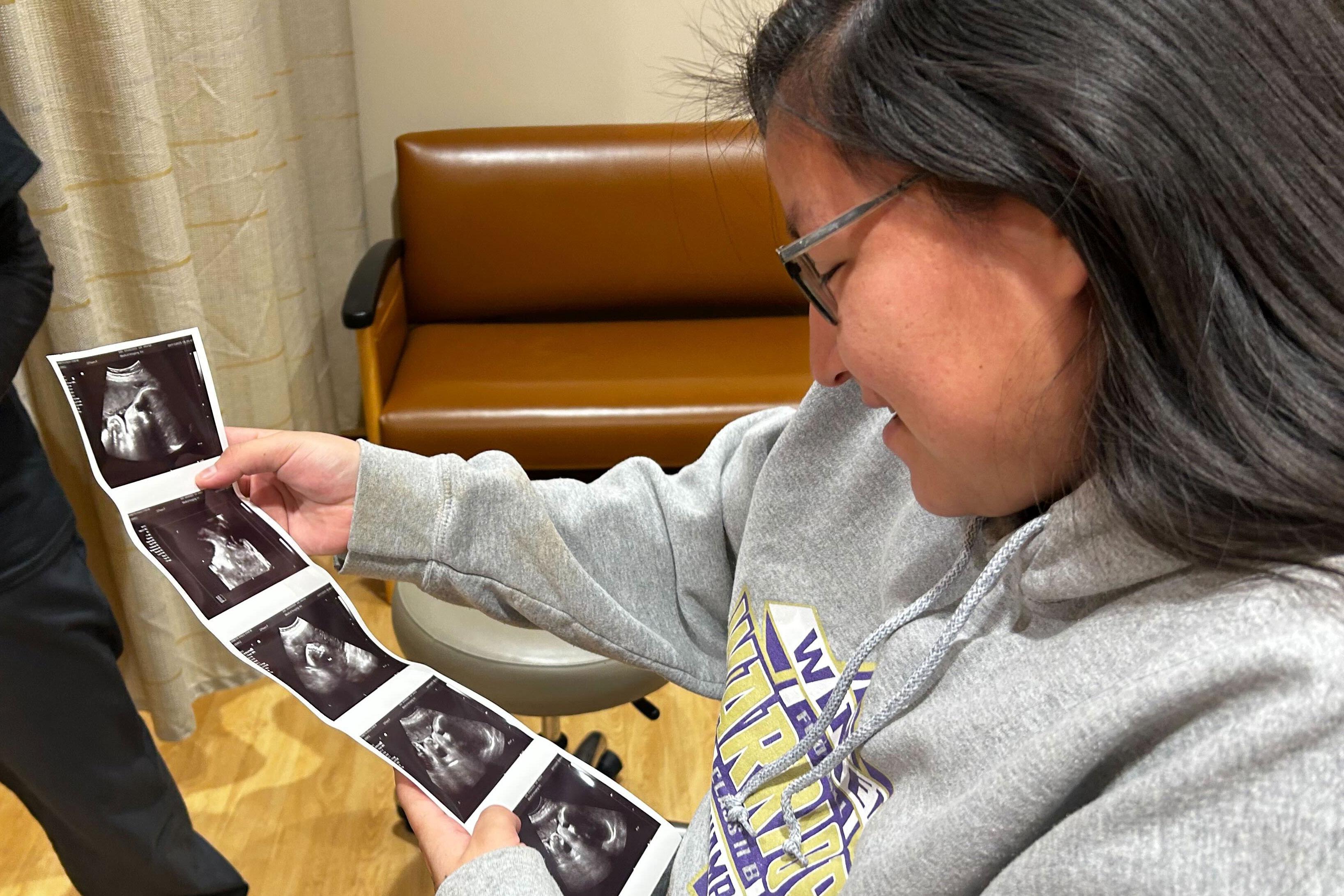 A woman in a gray sweatshirt holds a series of five ultrasound photos and smiles down at them.