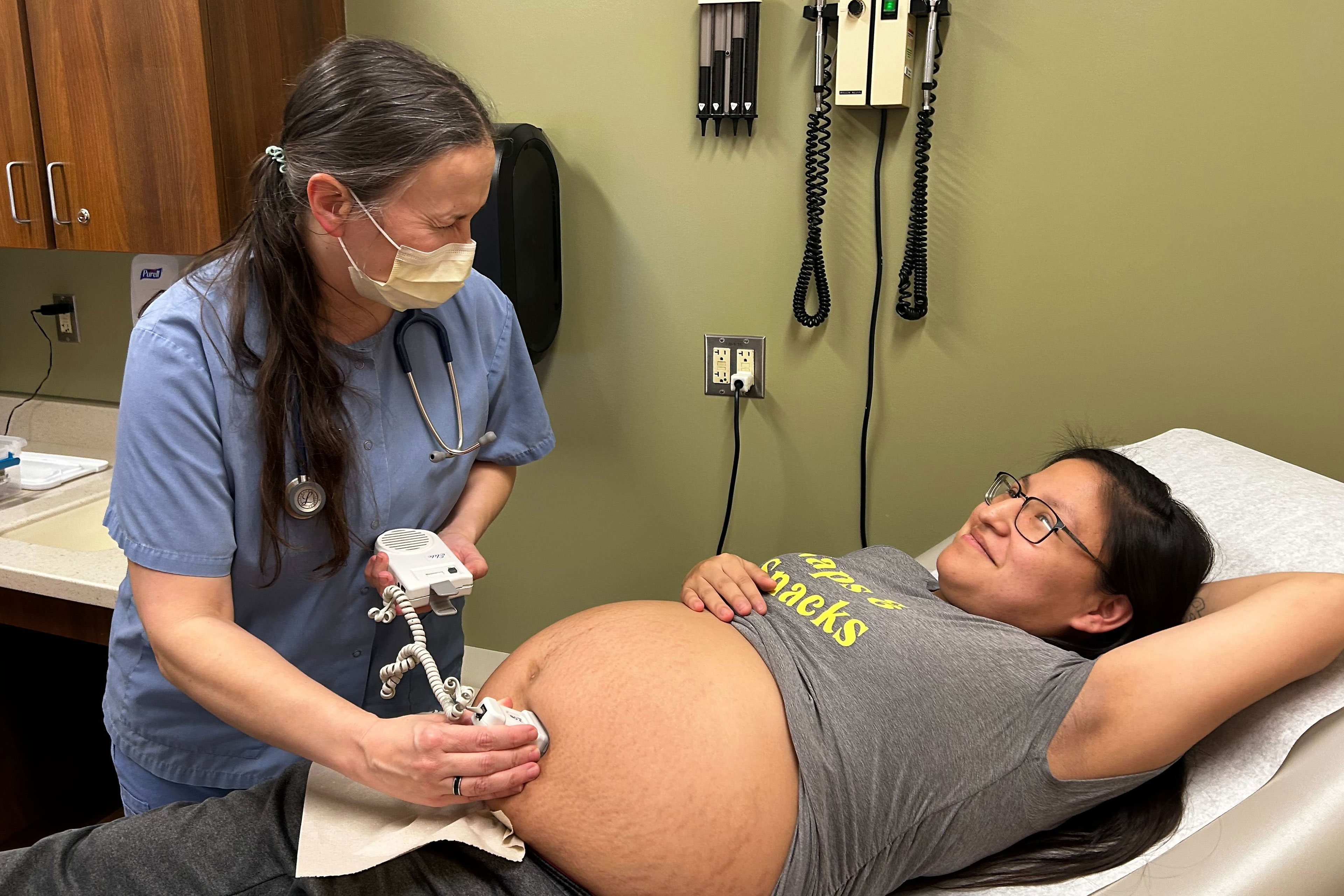 A woman in scrubs and a mask holds a device to the pregnant belly of a woman lying on her back on an exam table.