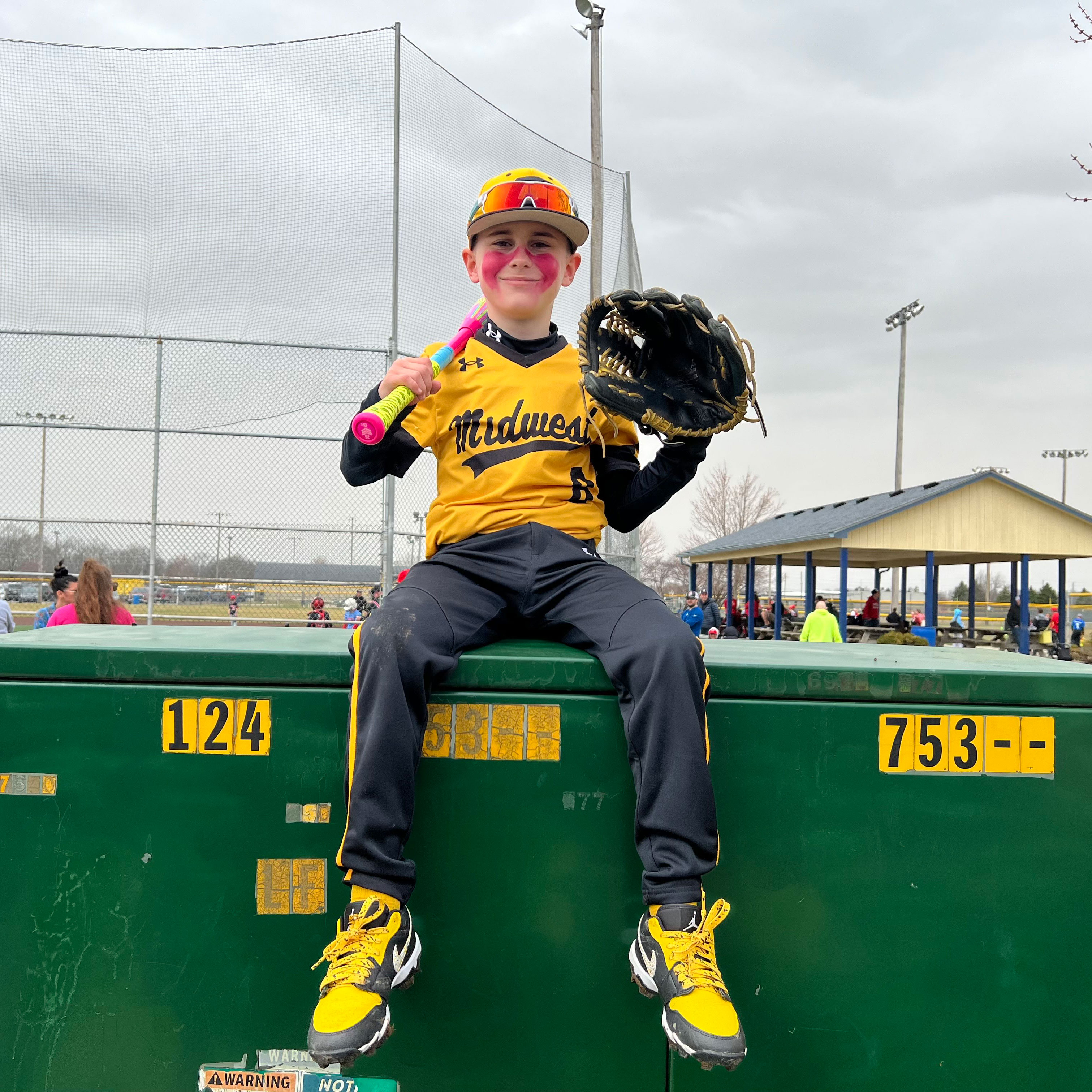 A boy in a baseball uniform and holding a bat and glove, sits atop a large green metal box with his legs dangling and smiles at the camera. A baseball field and covered seating area are behind him.