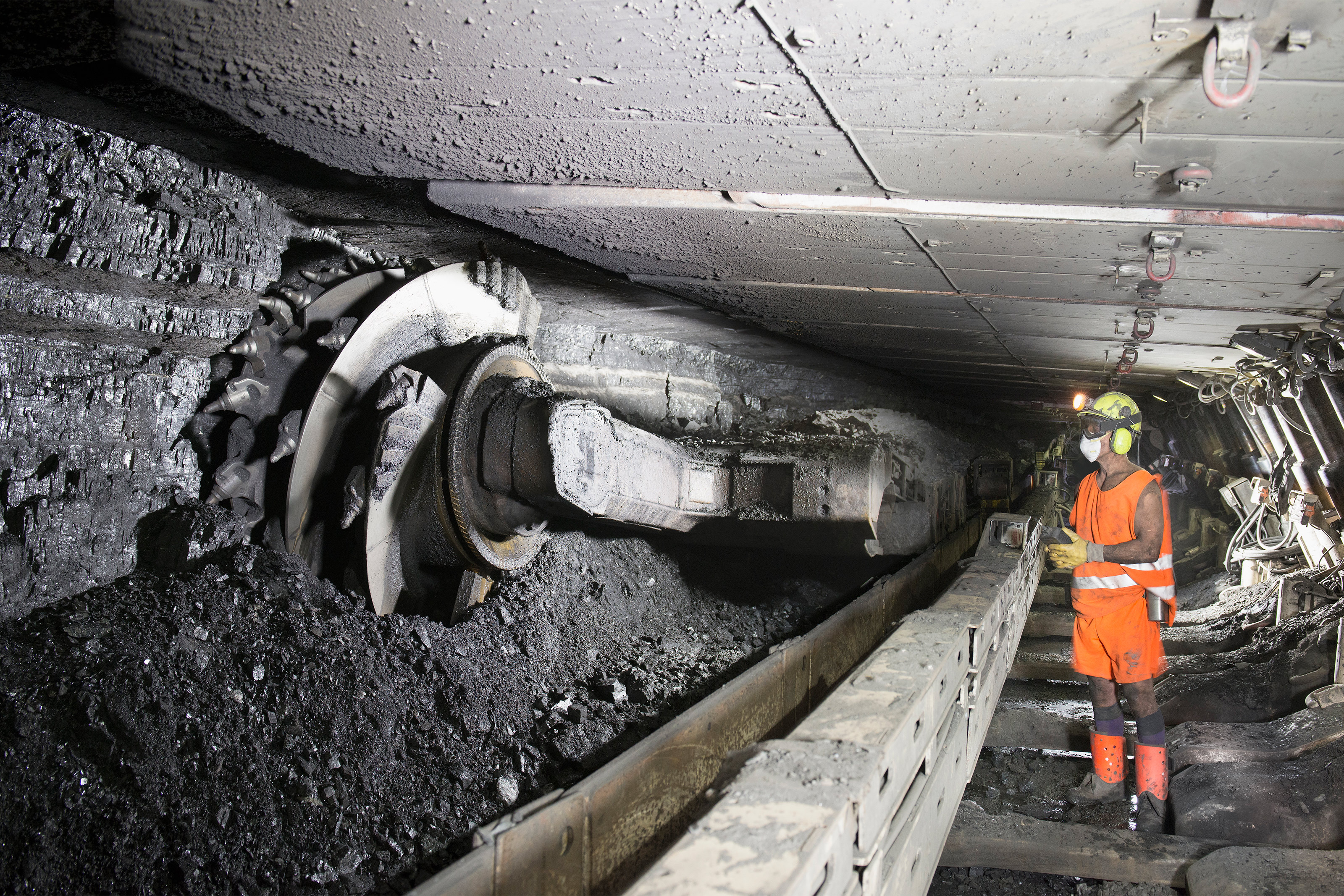 A photo of a worker wearing reflective gear, hard hat, and a mask, stands by a coal cutting machine in a coal mine.