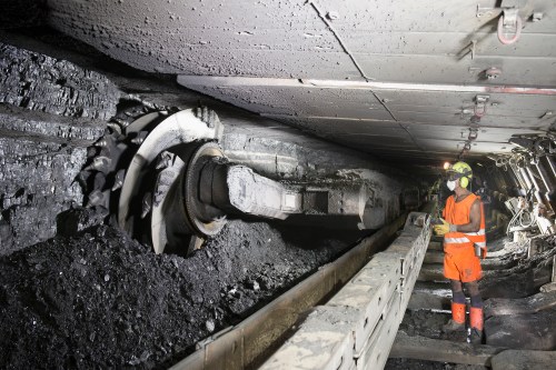 A photo of a worker wearing reflective gear, hard hat, and a mask, stands by a coal cutting machine in a coal mine.