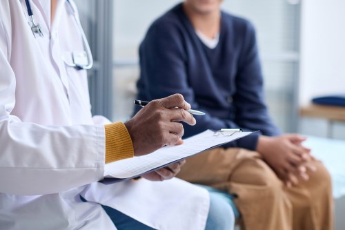 A photo of a doctor's appointment. A doctor reads off notes from a clipboard as a patient on the exam table listens.