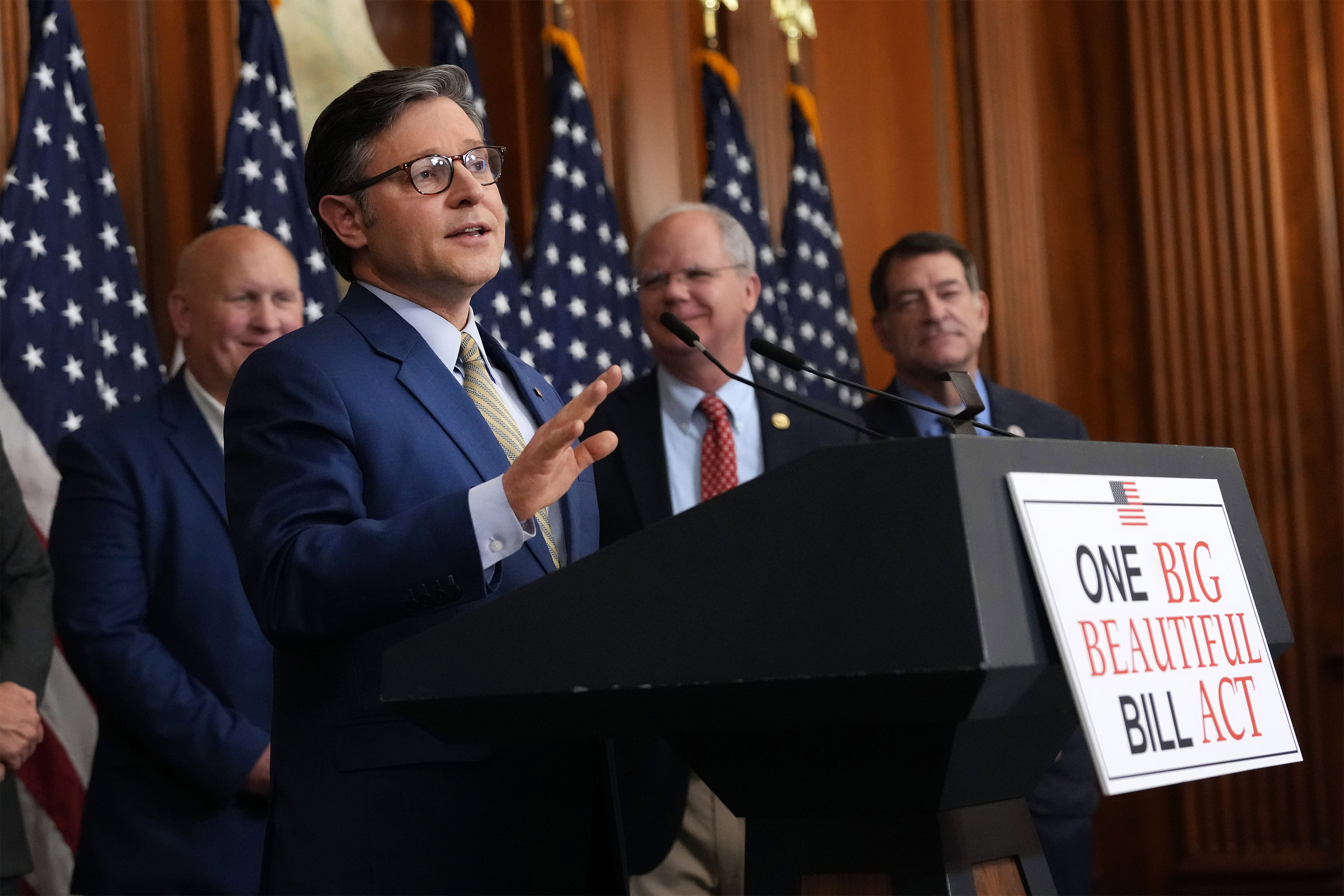 A photo of Mike Johnson speaking at a podium with a sign attached to it that reads "One Big Beautiful Bill Act."