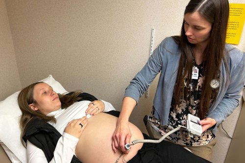 A woman in a white shirt and black vest pulled up to expose her belly lies on an exam table as another woman holds a device to the pregnant woman's stomach.