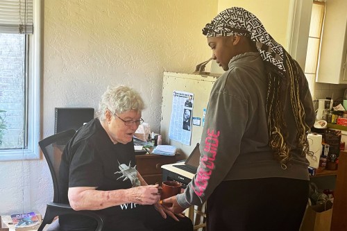 A photo of an aide bringing an elderly woman coffee at her desk.
