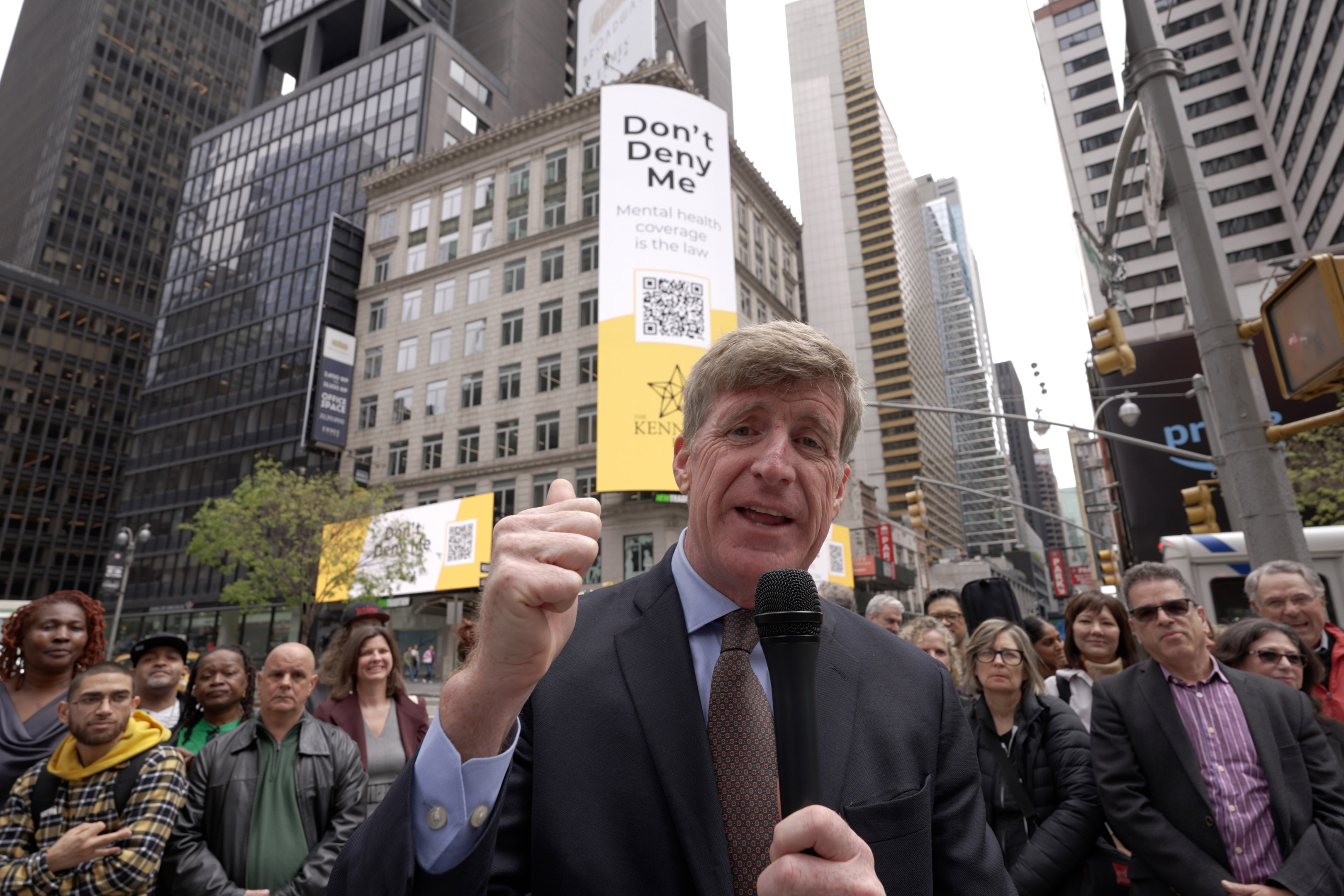 A photo of a man holding a microphone, speaking to the camera, with a crowd behind him.