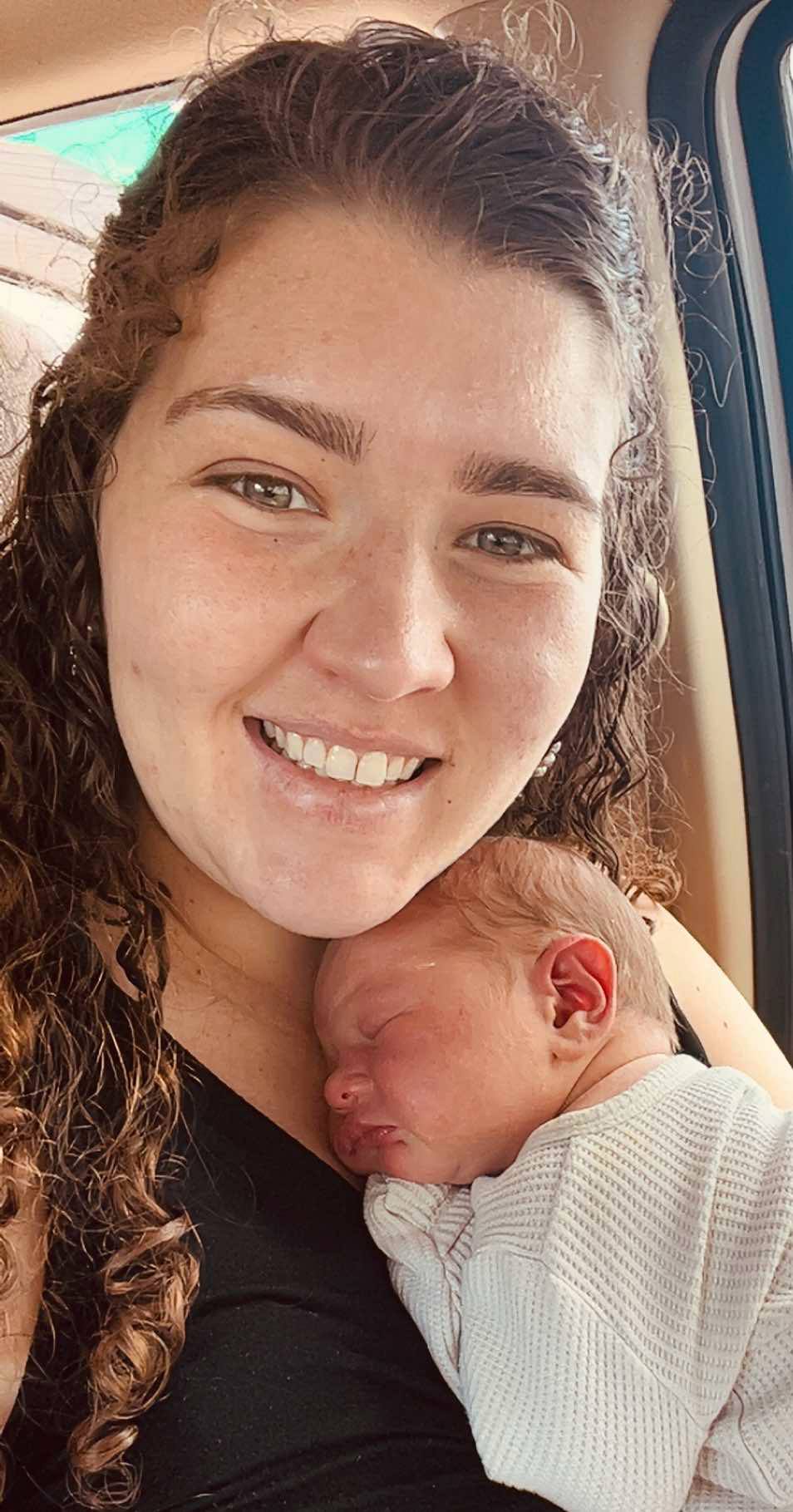 In this selfie, a woman with long curly hair holds a young baby to her chest and smiles at the camera.