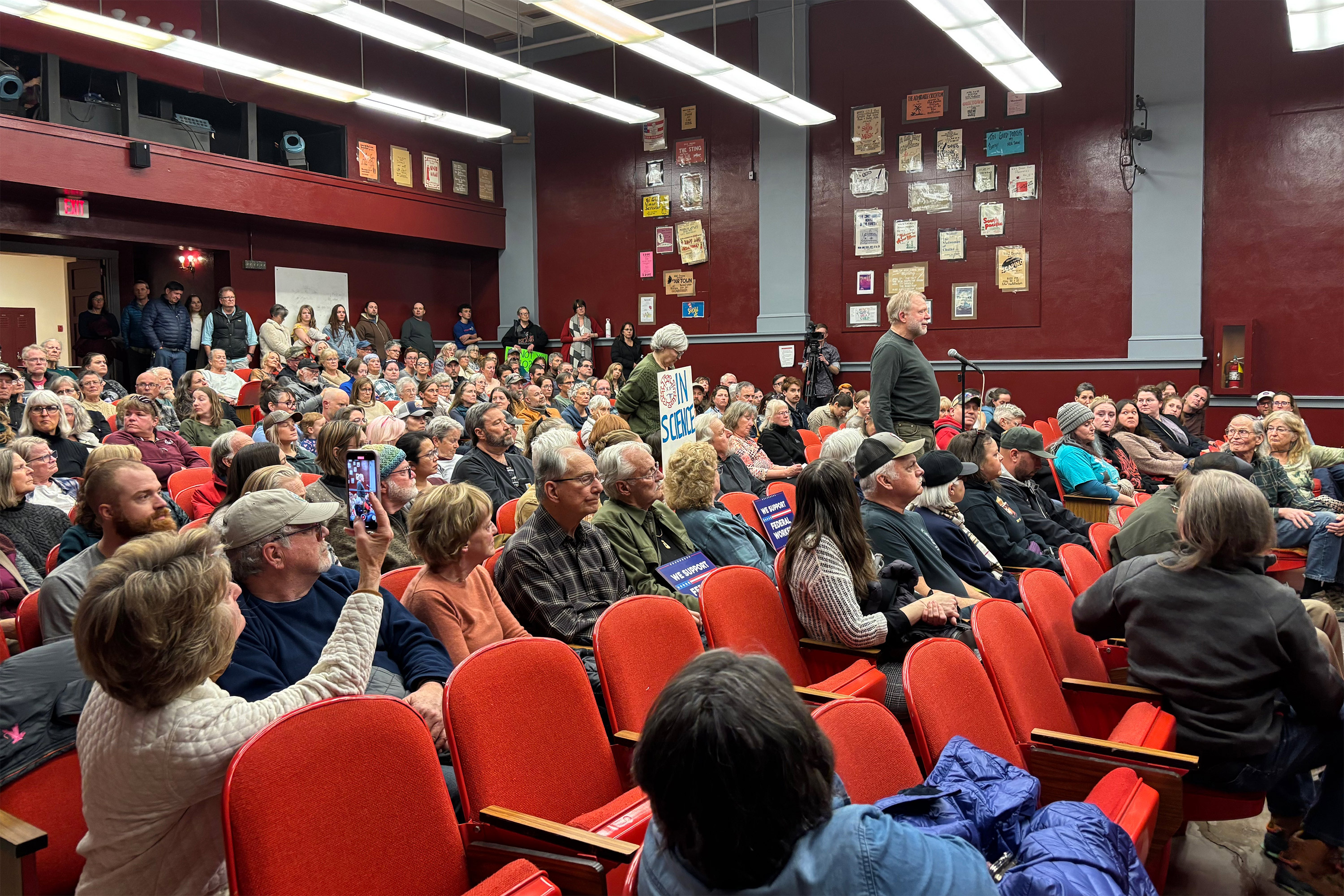 A photo of a school auditorium filled with protesters. A man speaks at a microphone. Others in the room hold signs or film him on their phones.