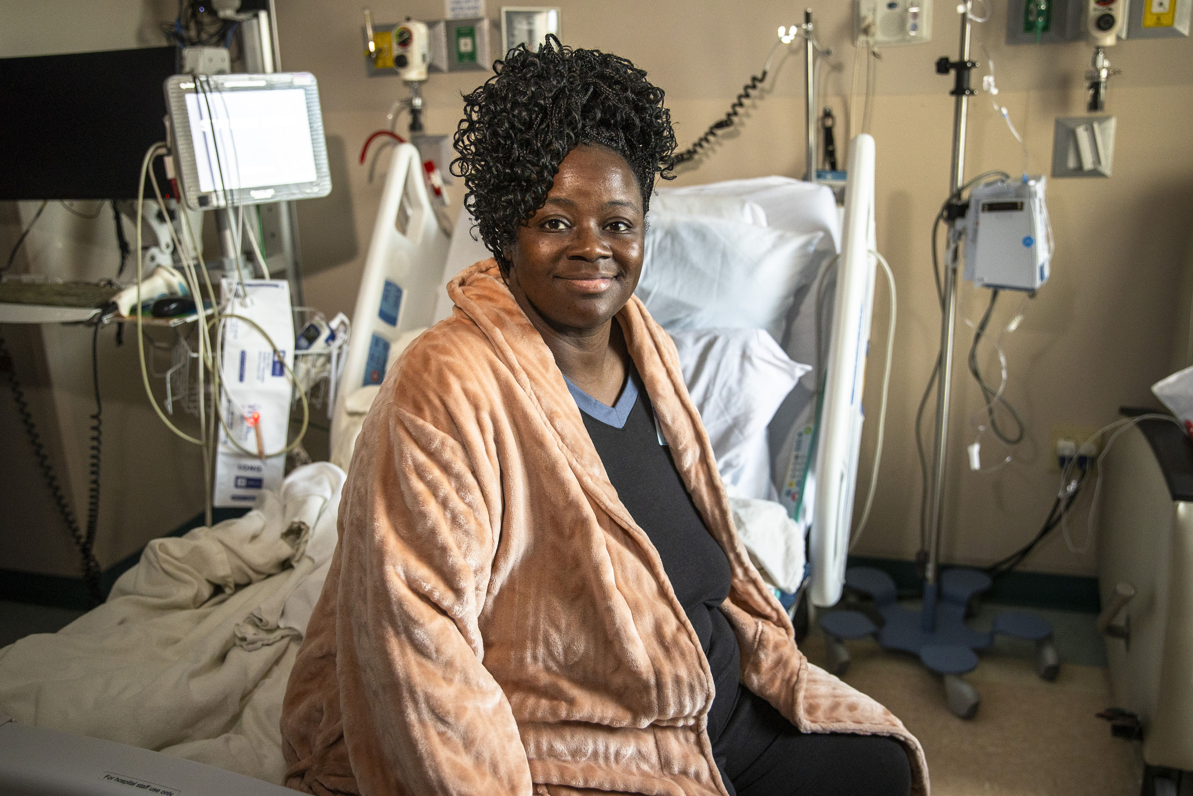 A photo of a woman sitting on a hospital bed with her legs off to the side. She smiles, facing the camera.