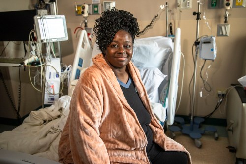 A photo of a woman sitting on a hospital bed with her legs off to the side. She smiles, facing the camera.
