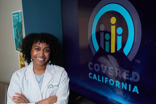 A photo of a woman in a white lab coat posing by a screen with the Covered California logo.