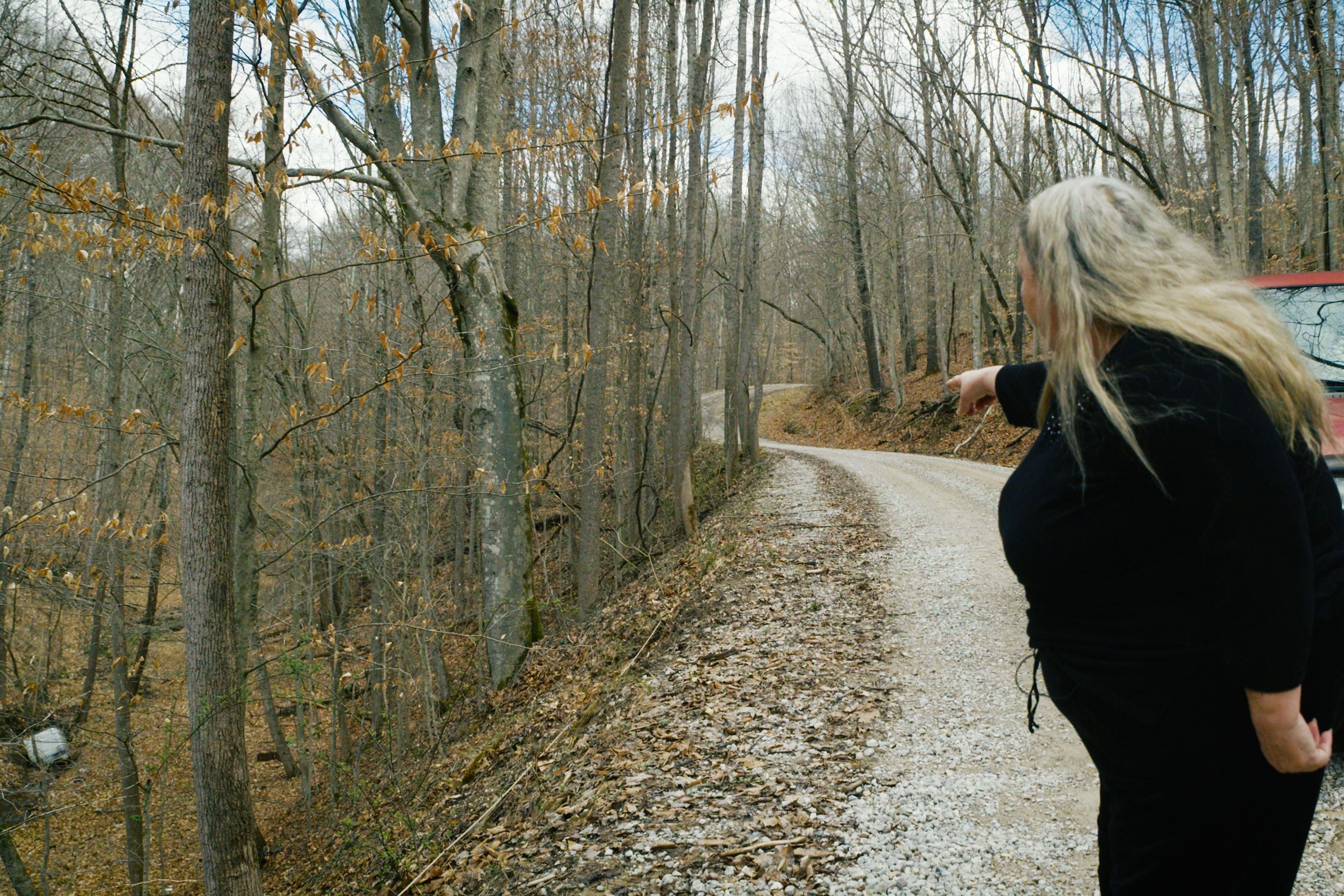 A woman stands on a gravel tree-lined road and points to a wire on the ground