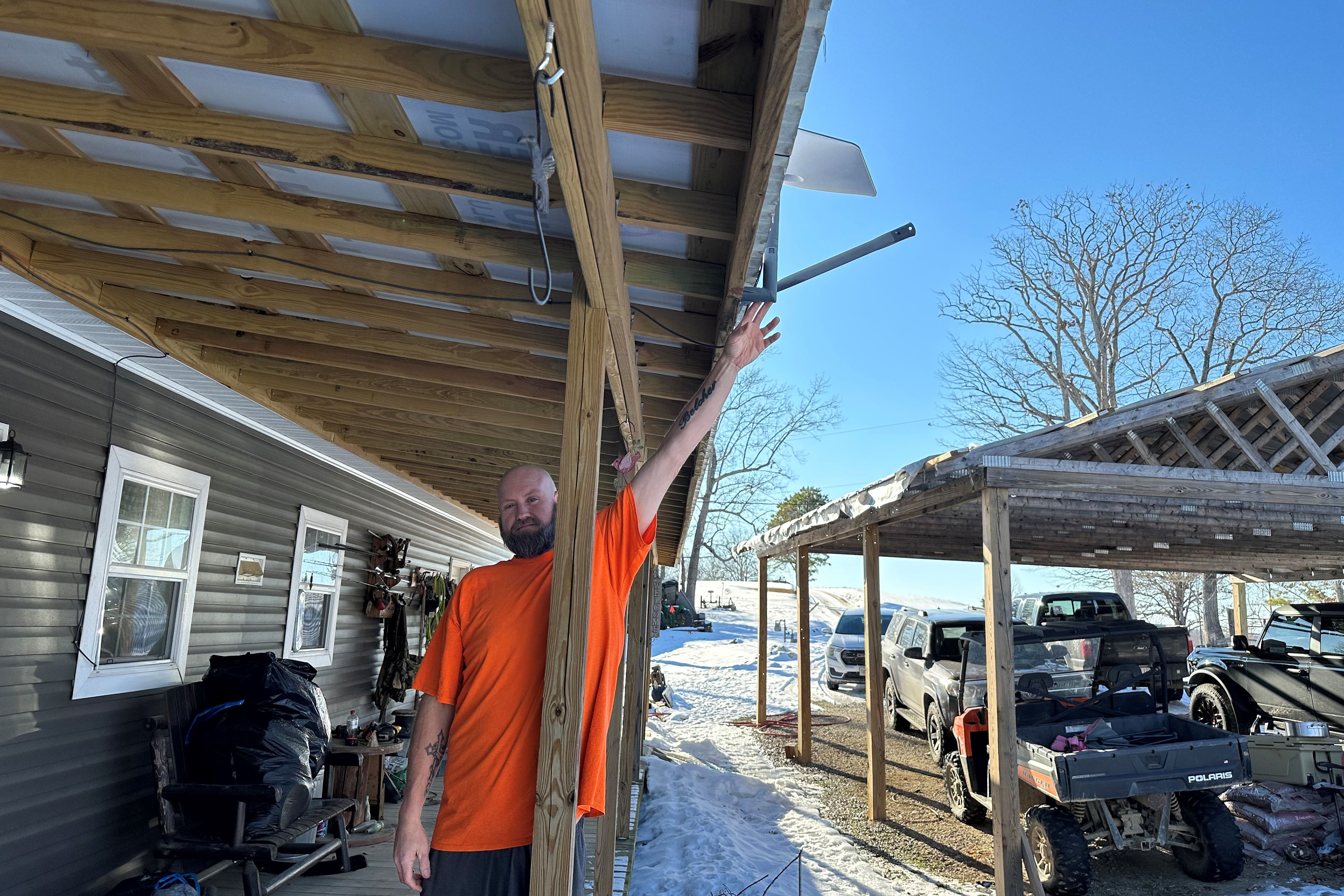 A man in an orange shirt stands on a porch and points to a satellite antenna